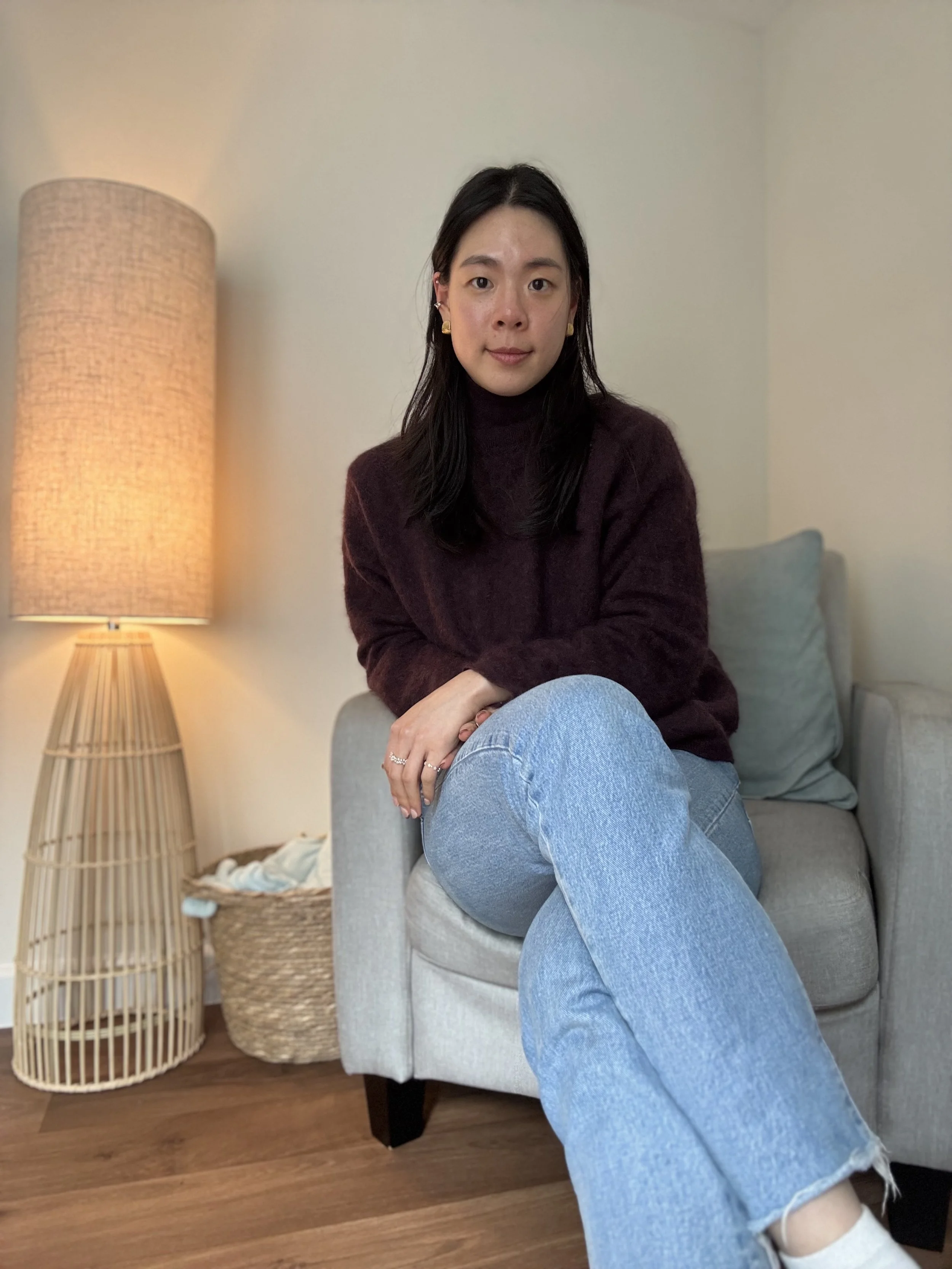 A woman with long dark hair sitting on a light-colored armchair in a cozy room. She wears a maroon sweater and light blue jeans. The background features a lit floor lamp, a basket with laundry, and a cushion on the armchair.