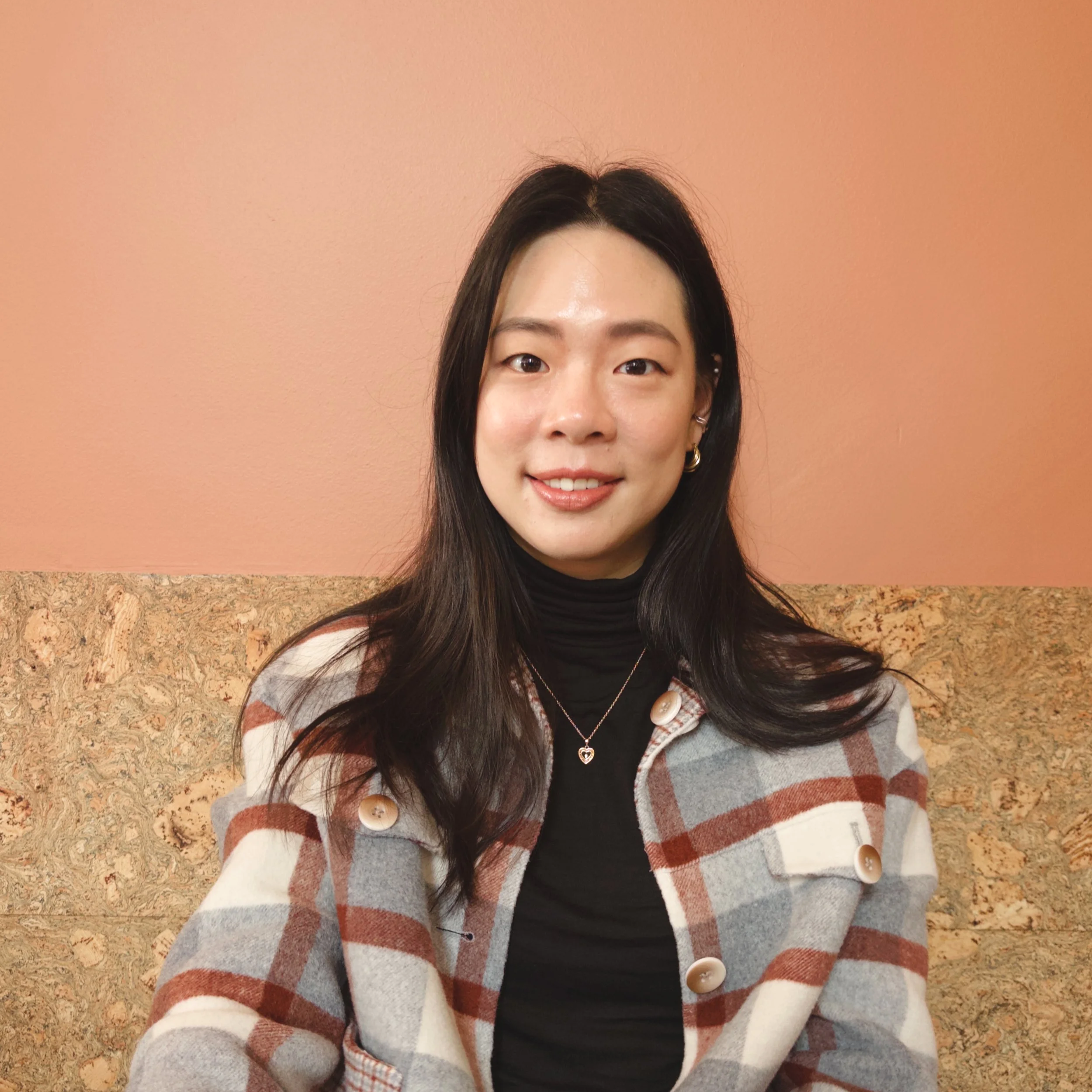 A woman with dark hair smiling, wearing a black turtleneck, heart-shaped necklace, and a plaid coat with large buttons, standing in front of a pink wall with a granite floor.