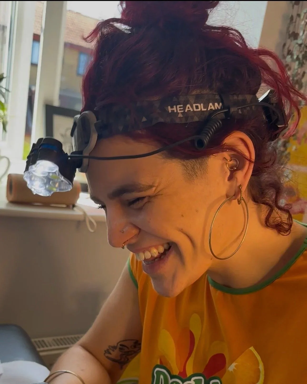 A woman with curly red hair wearing a head torch with a bright LED light, earrings, and a yellow shirt, smiling and looking downward.