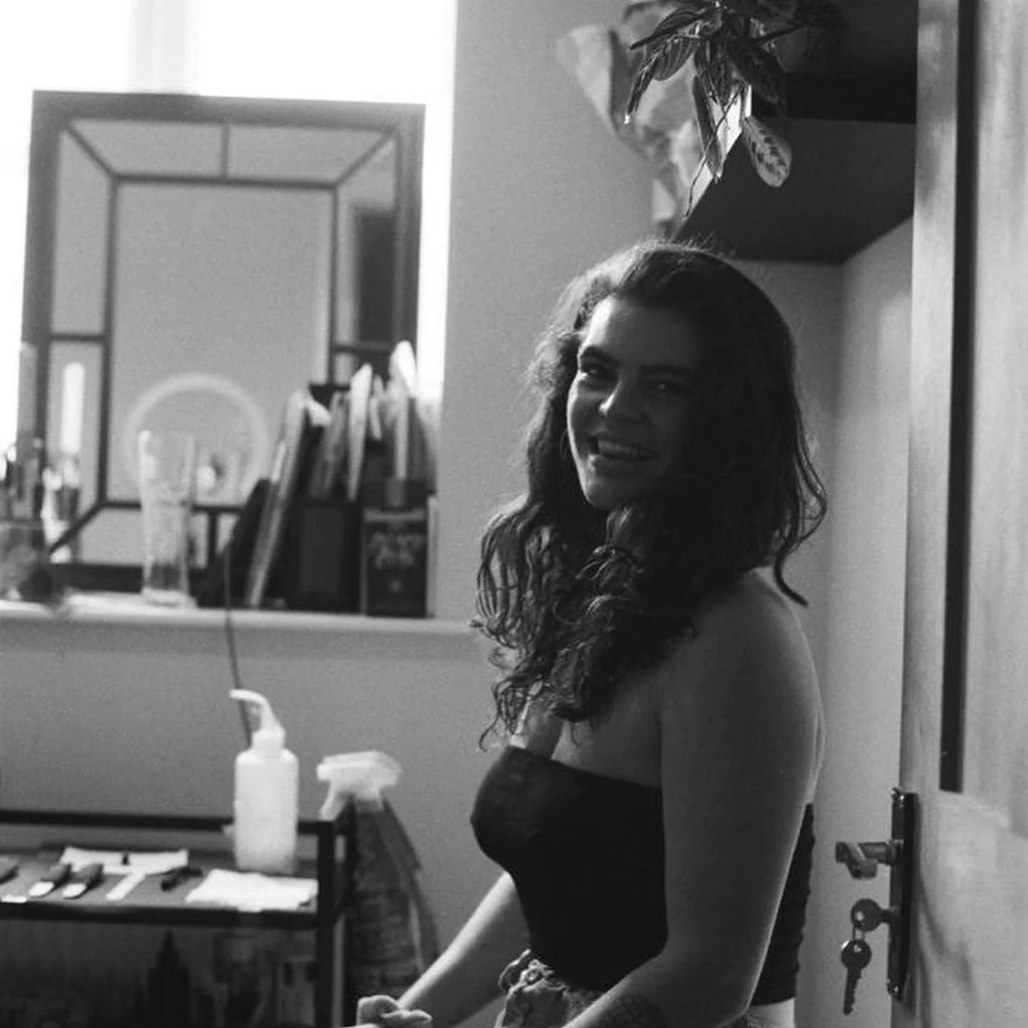 A smiling woman with long, curly hair, wearing a strapless top, standing in a room with shelves, tattoo bottles, and a mirror.