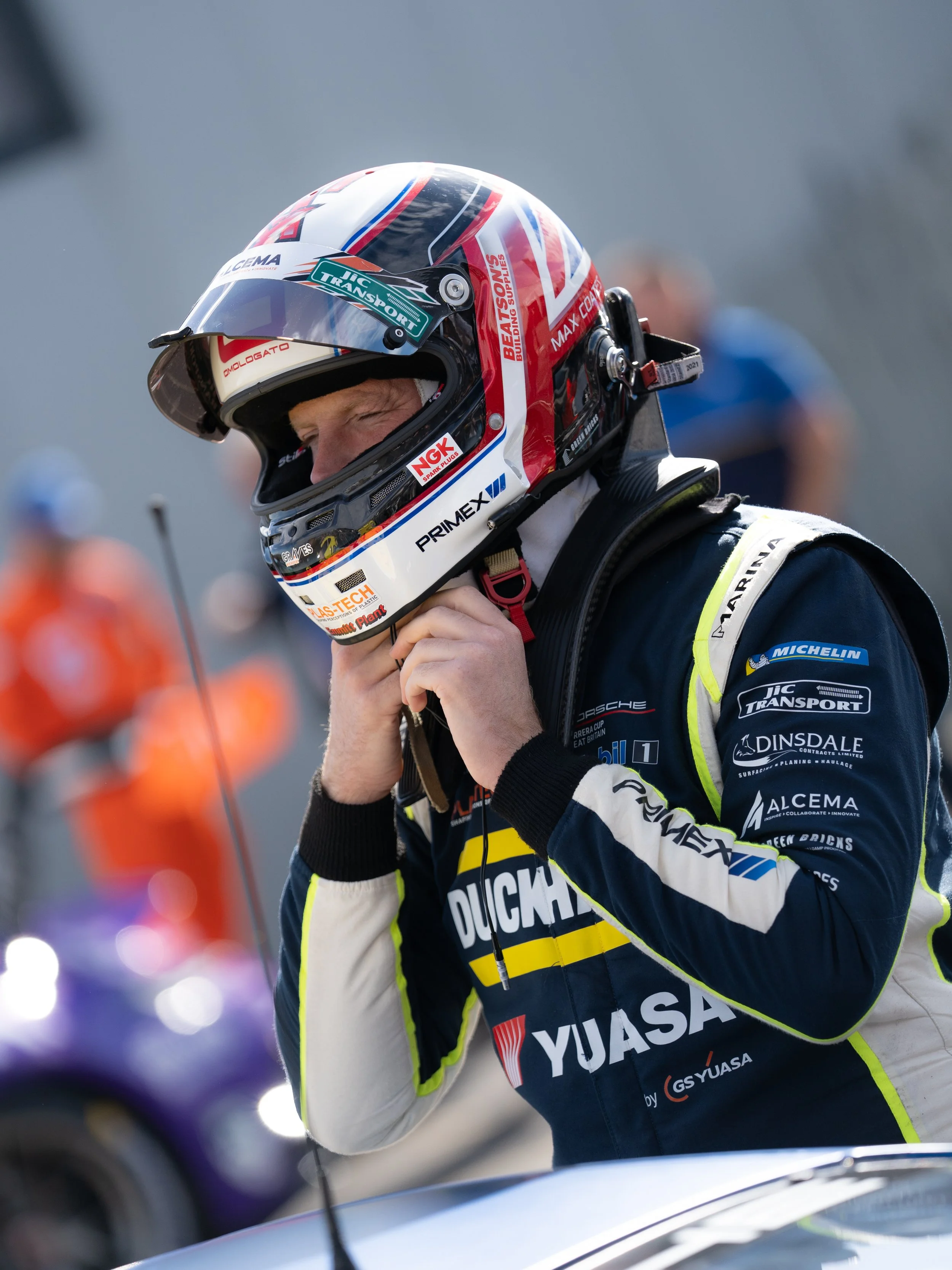 Race car driver adjusting helmet strap before a race, wearing a black racing suit with yellow and white accents and various sponsor logos.