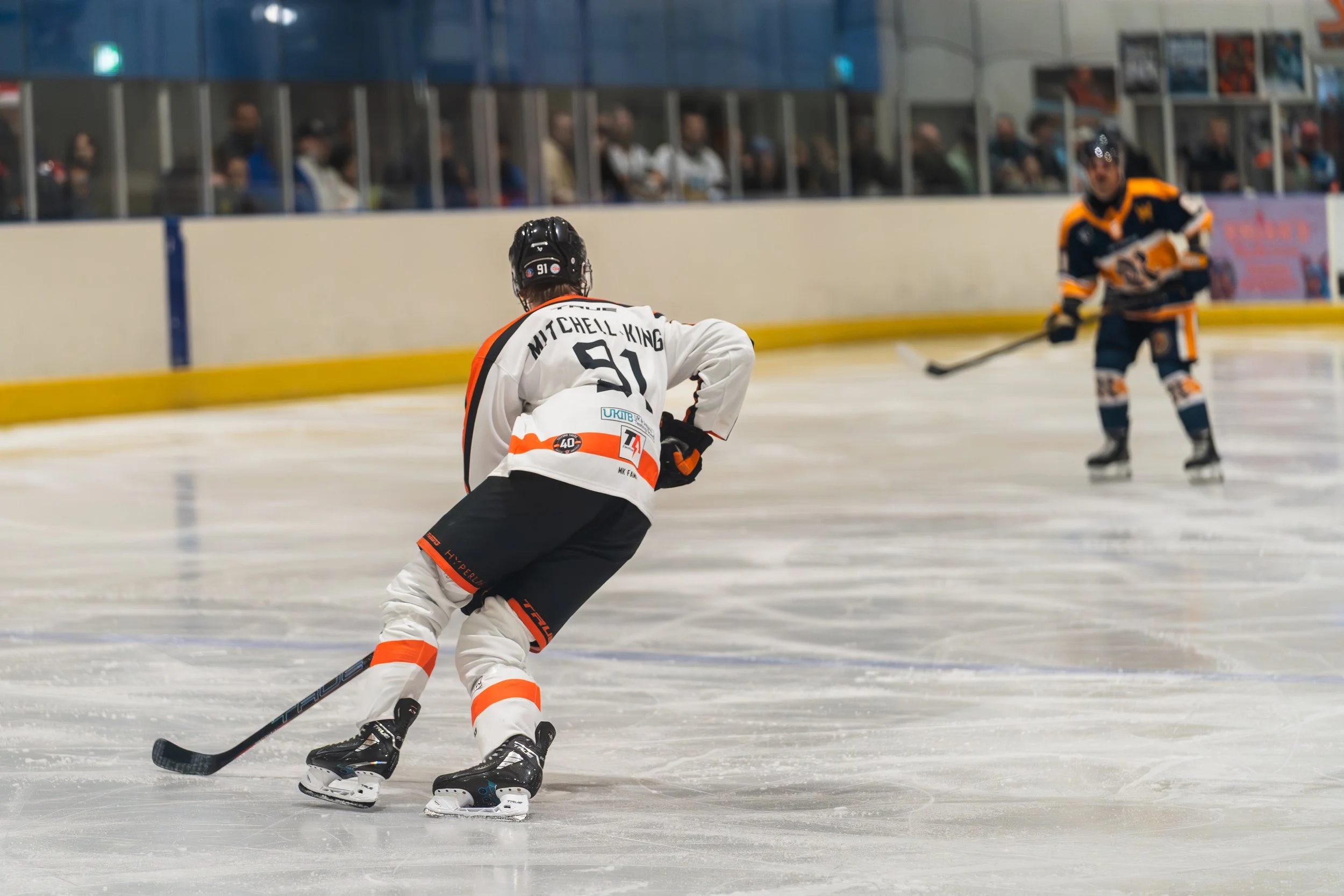 Hockey game with two players on ice, one in white and black jersey with number 91, and the other in a dark jersey with orange and white, seen from behind.