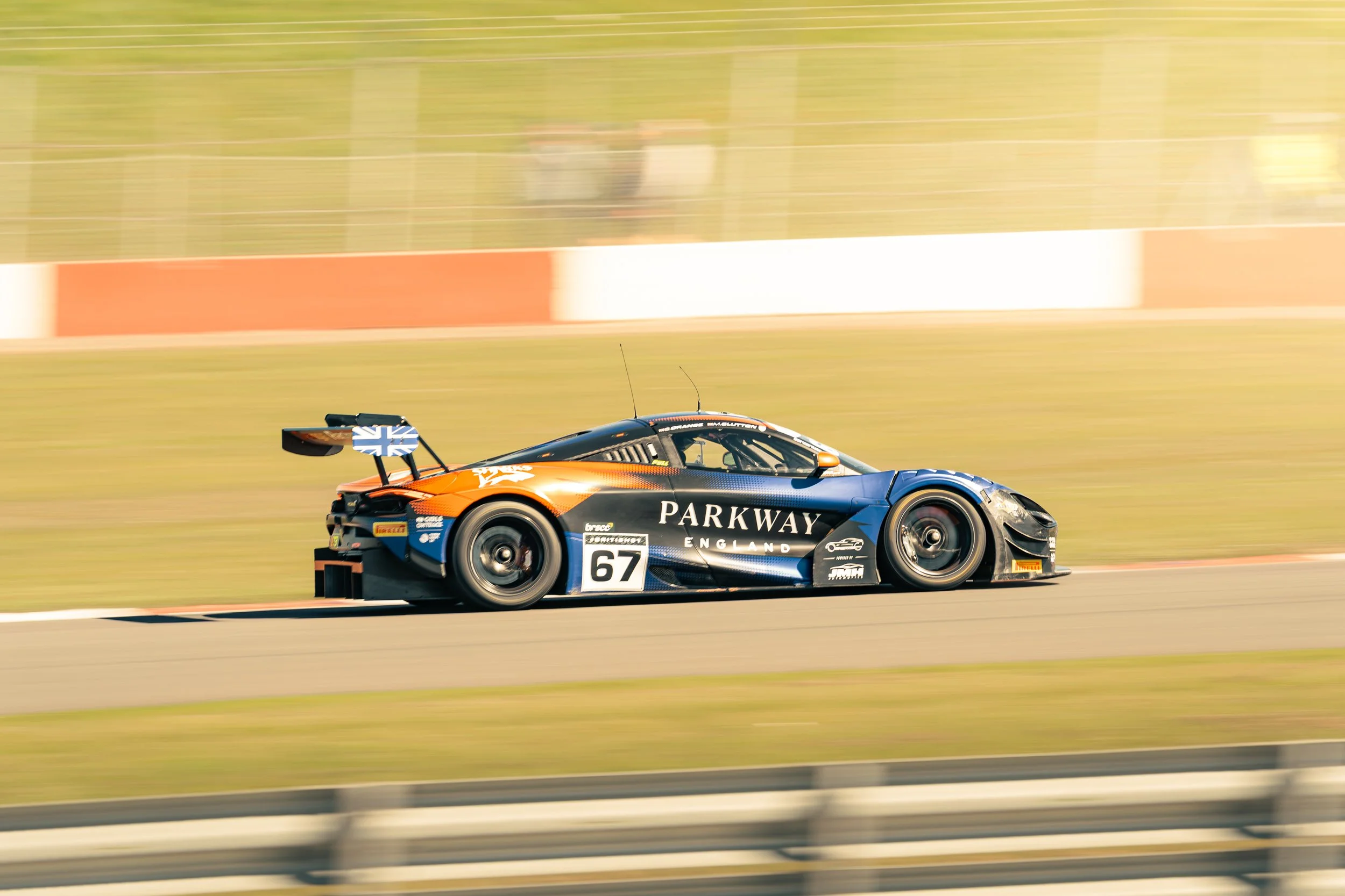 A race car on a track with a blue and orange design, the number 67, and sponsorship logos, moving at high speed.