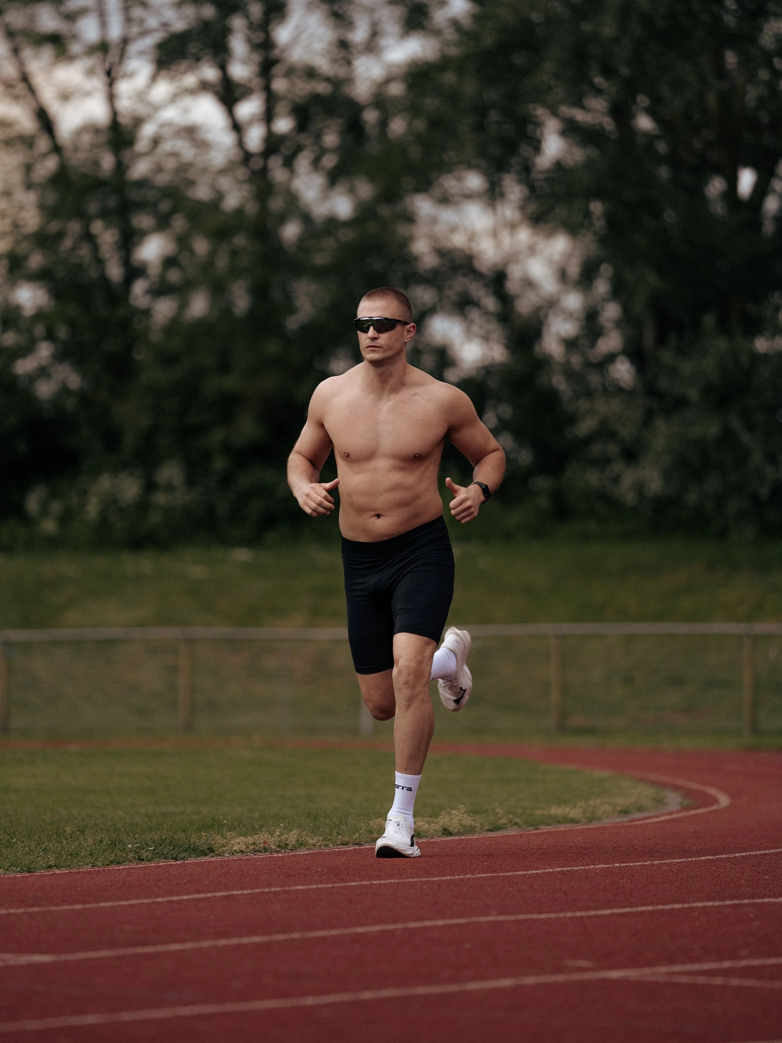 A shirtless man running outdoors on a track, wearing black shorts, white running shoes, sunglasses, and a sports watch, with trees in the background during the evening.
