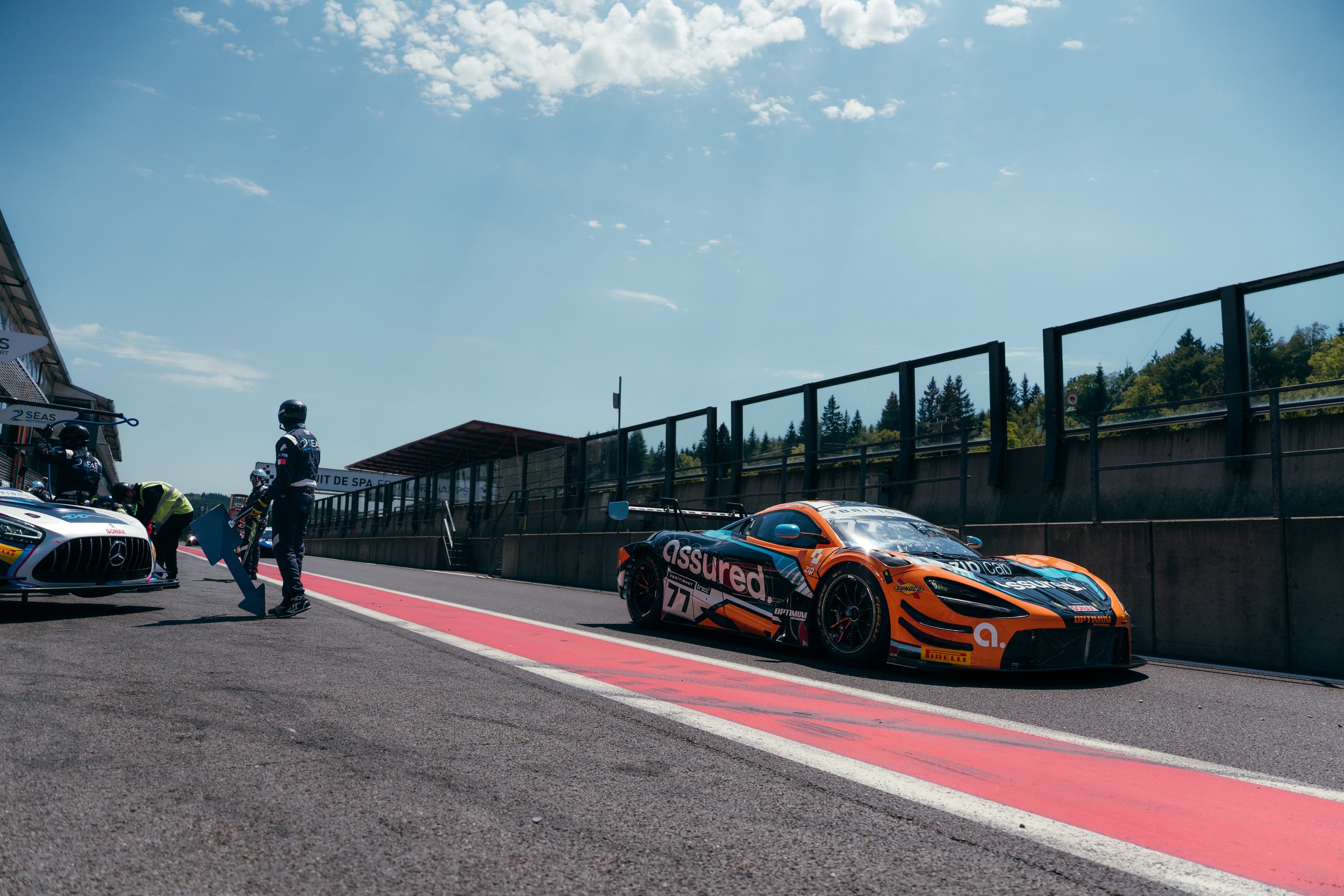 Race cars and crew members preparing in the pit lane at a motorsport race track.