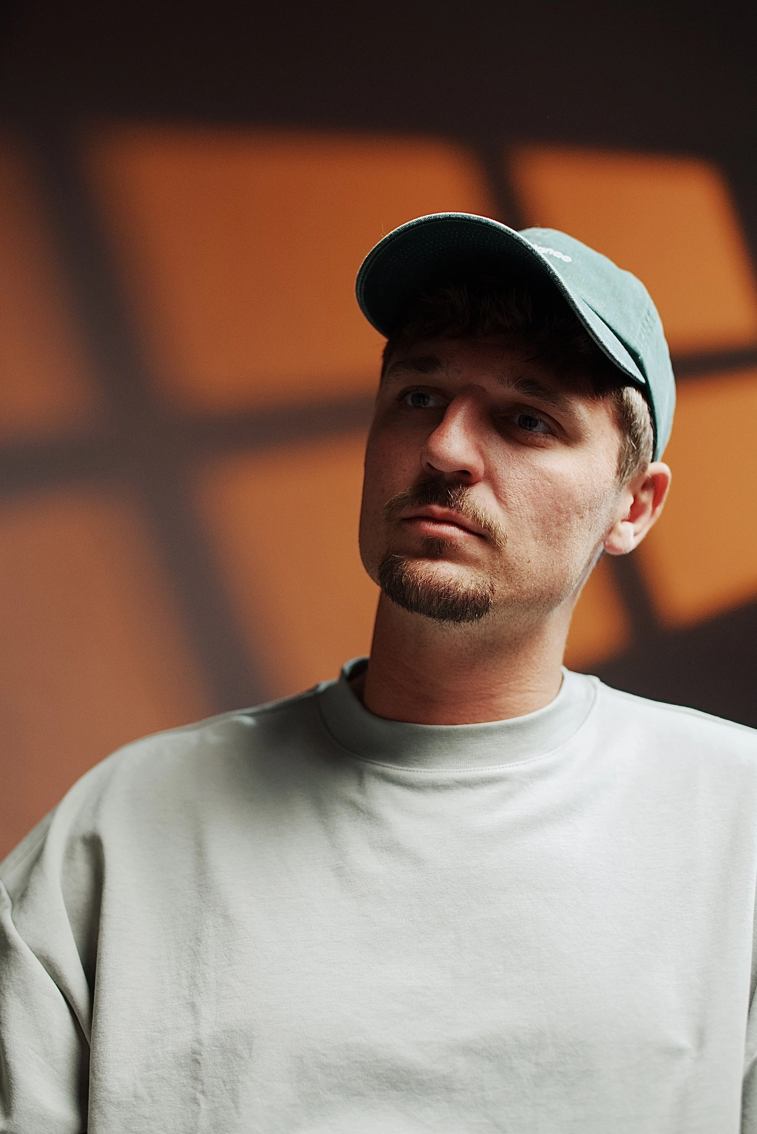 Close-up of a young man with a beard and mustache, wearing a grey sweatshirt and a teal cap, looking to the side against a blurred orange and black background.