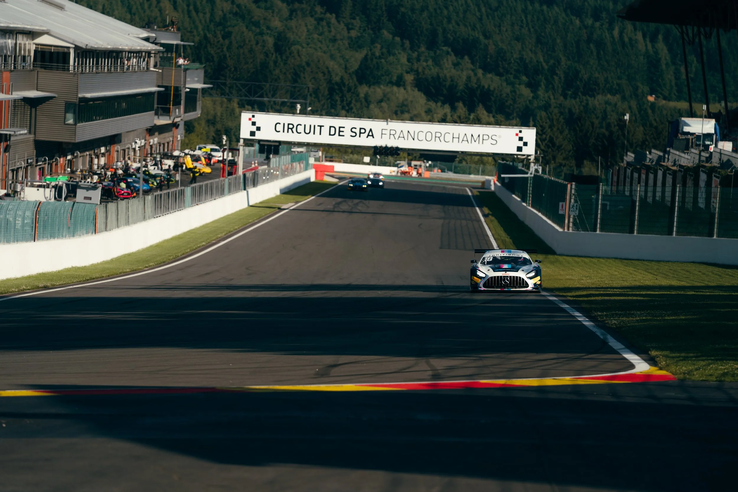 Race car on a track with a sign reading 'Circuit de Spa Francorchamps,' crowd in the stands, and forested hills in the background.