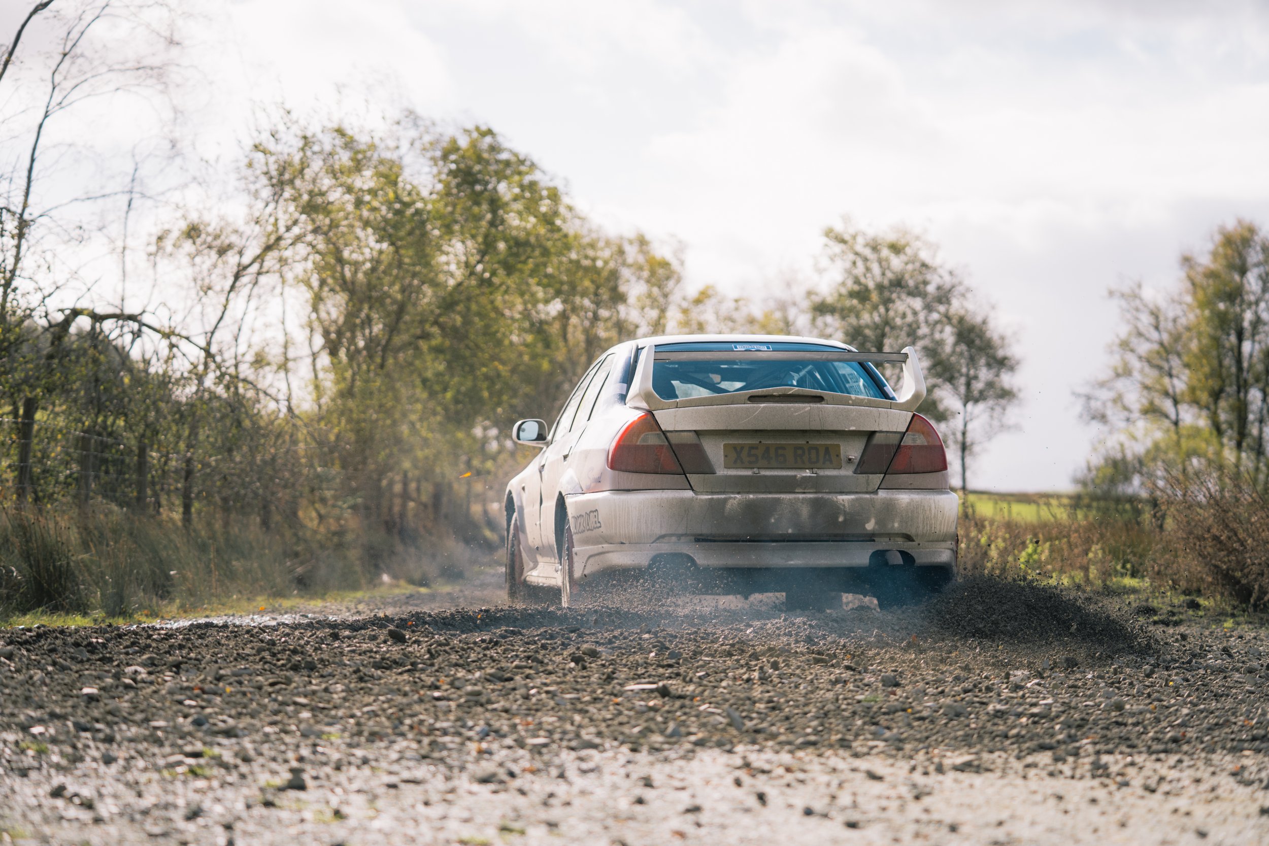 A rally car driving on a gravel road with dust and dirt kicked up behind it, surrounded by trees and a cloudy sky.