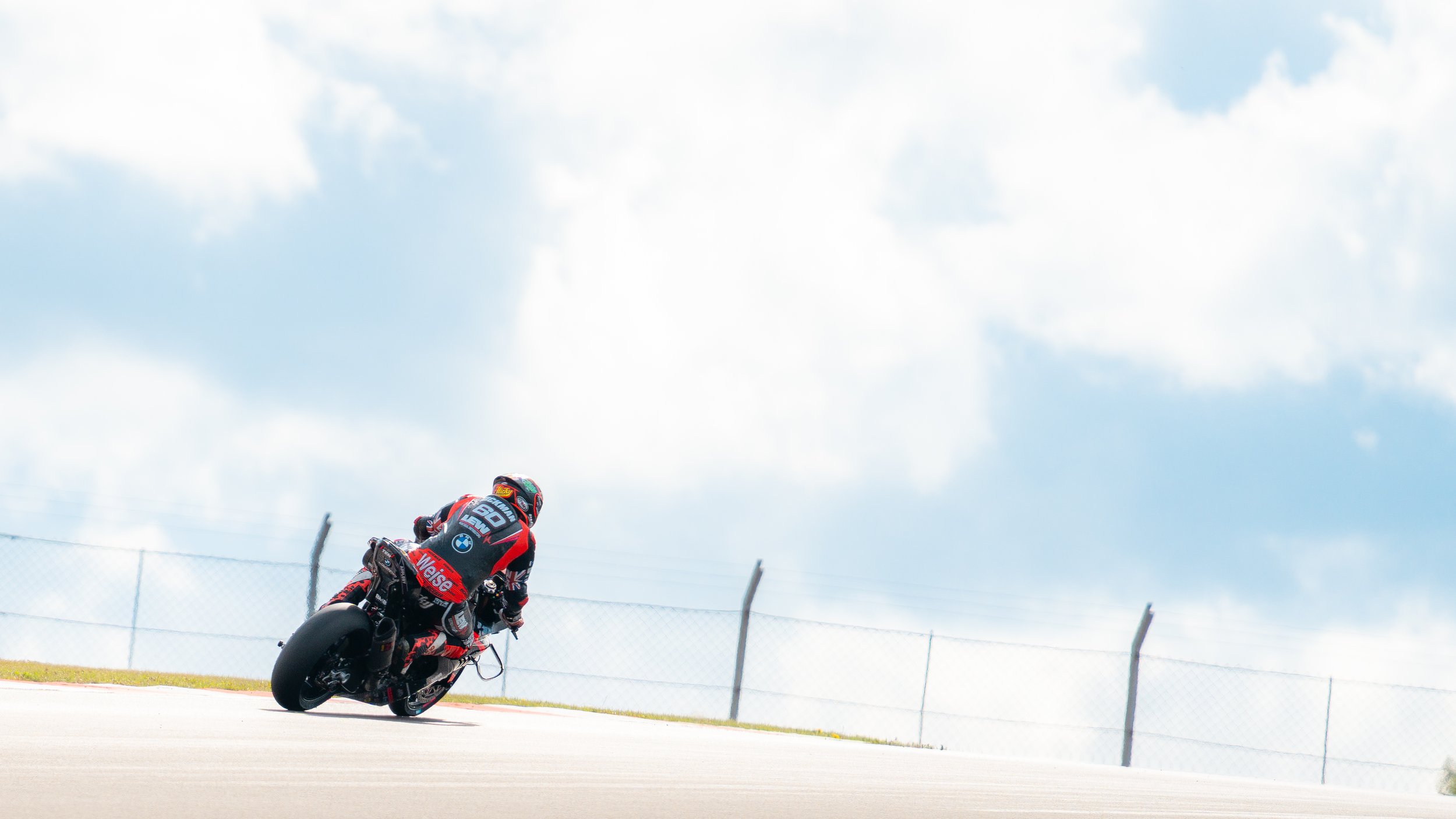 A motorcycle racer in a black and red racing suit leaning into a turn on a racetrack, with a chain-link fence and sky with clouds in the background.