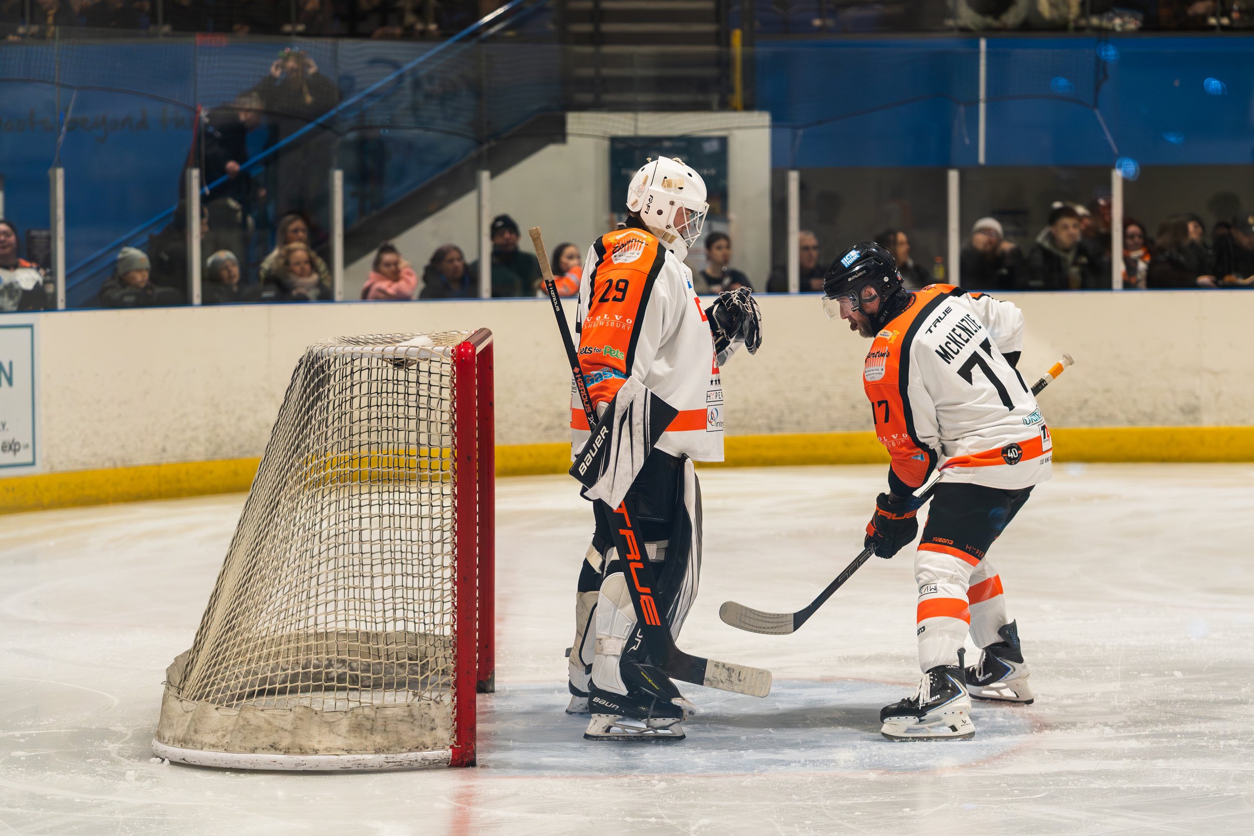 Two ice hockey players near the goal post, one dressed as a goalie with white gear and the other player in a white and orange jersey, on the ice rink during a game, with spectators in the background.