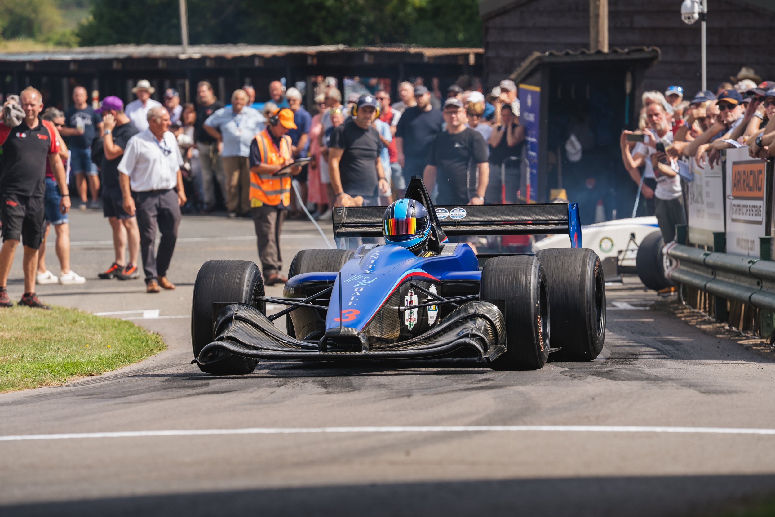 A blue Formula racing car with the number 3 is on the track, surrounded by a crowd of spectators and officials. The driver is wearing a helmet, and there's a hill with trees in the background.