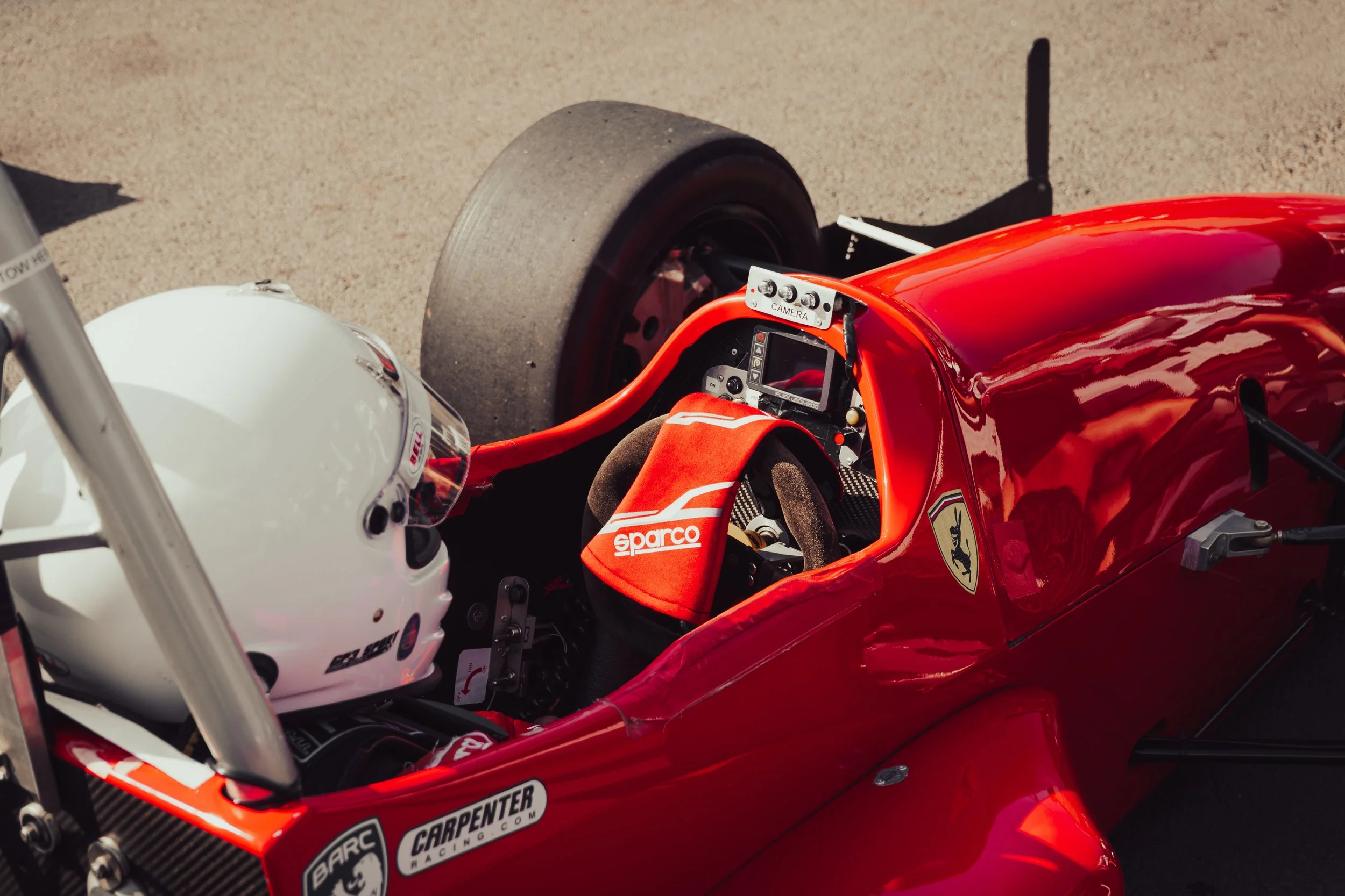 A red racing car with a white driver helmet inside, featuring various sponsor logos, including Sparco. The car is positioned on a sandy surface, with black tires and a detailed interior, including a dashboard and steering wheel.