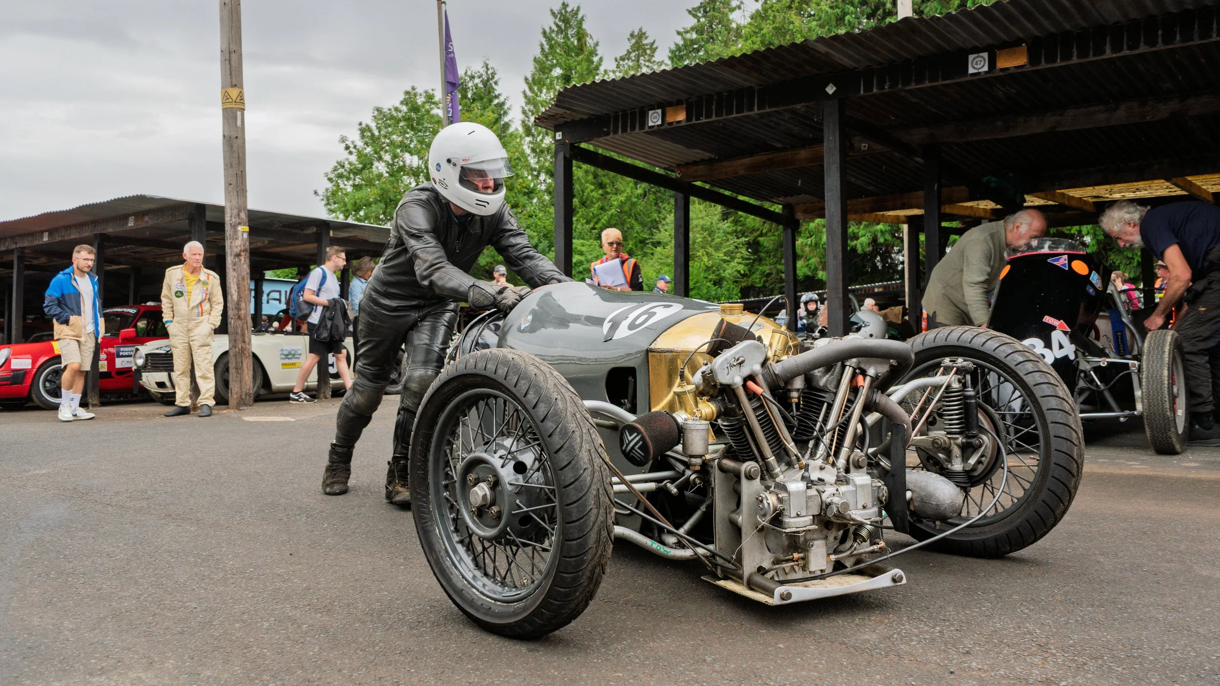 A person wearing a white helmet and black leather suit pushes a vintage racing car with an exposed engine and black tires, in a paddock area with other vintage cars and people in the background.