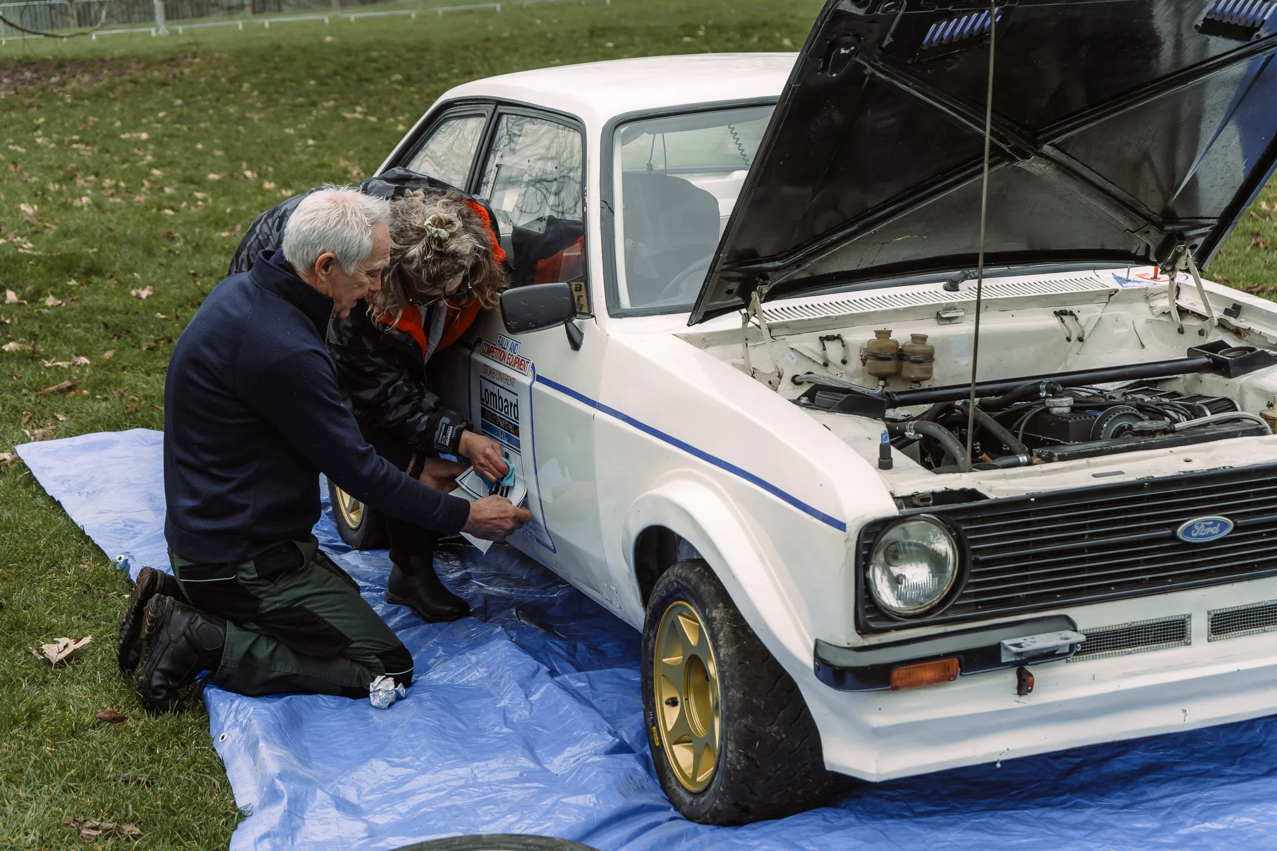 Mechanics servicing a classic Ford Escort rally car with the bonnet open at the Lombard Rally Festival
