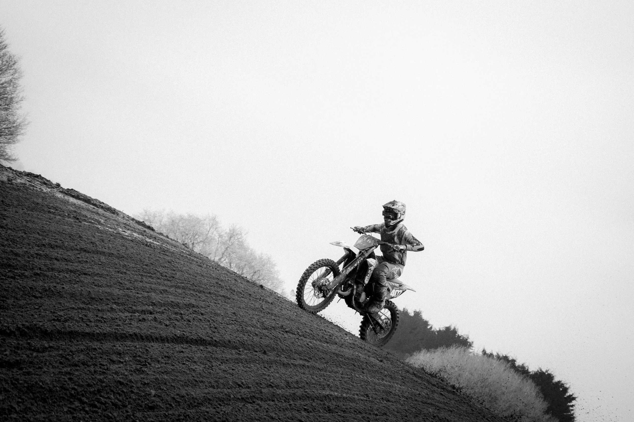 A person riding a dirt bike up a steep hill on a motocross track, wearing protective gear including a helmet, goggles, and a jacket.