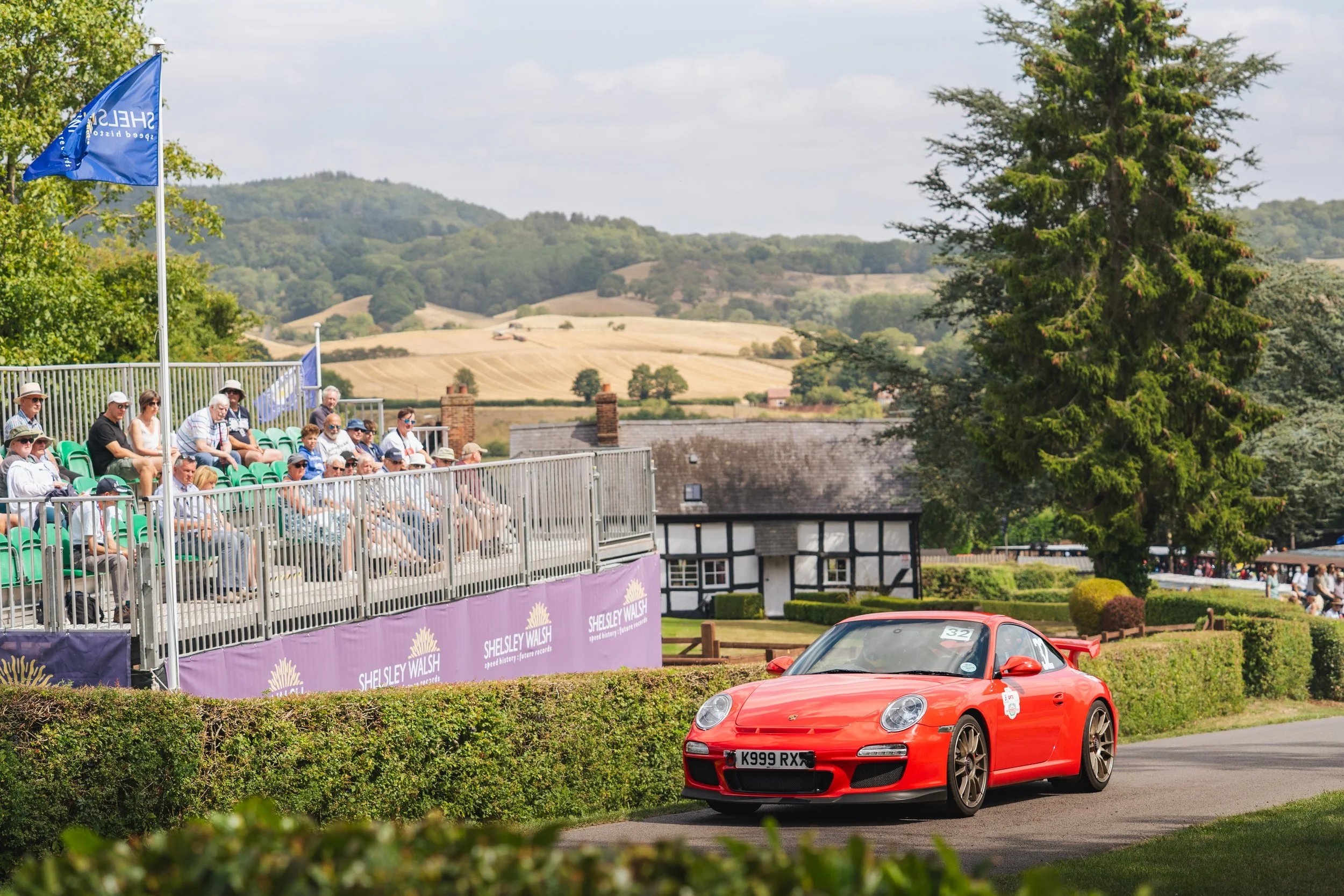 A red Porsche sports car racing on a track with spectators watching from the stands and a scenic countryside in the background.