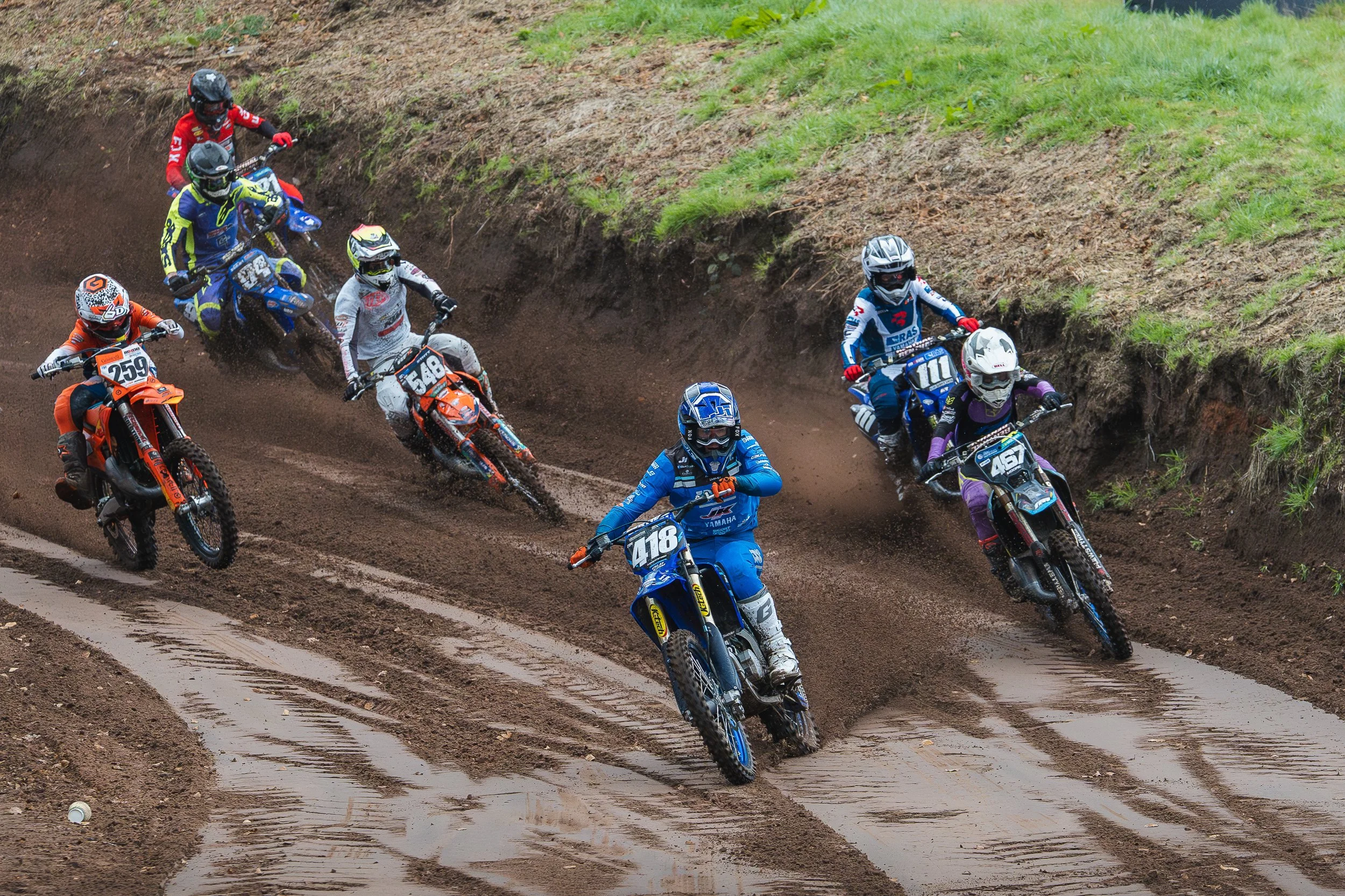 Pack of motocross riders cornering through a muddy berm at the British Motocross Championship Hawkstone International