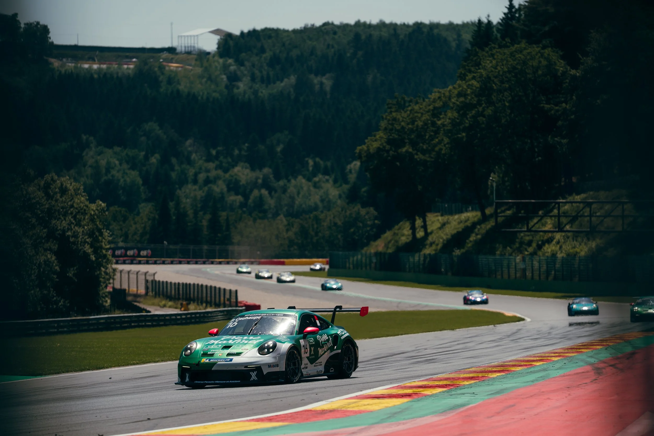 A green and white race car on a track with several other cars in the background, surrounded by green hills and trees, during a race.