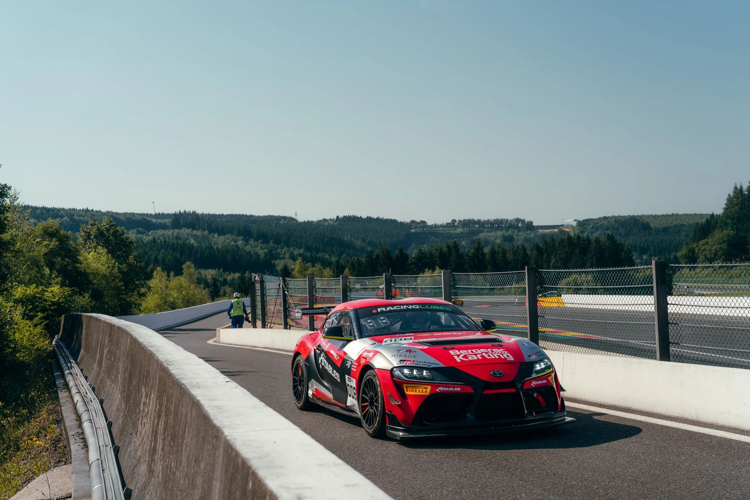 A red and black race car on a race track with a driver inside, surrounded by a barrier wall, with green trees and hills in the background during daytime.