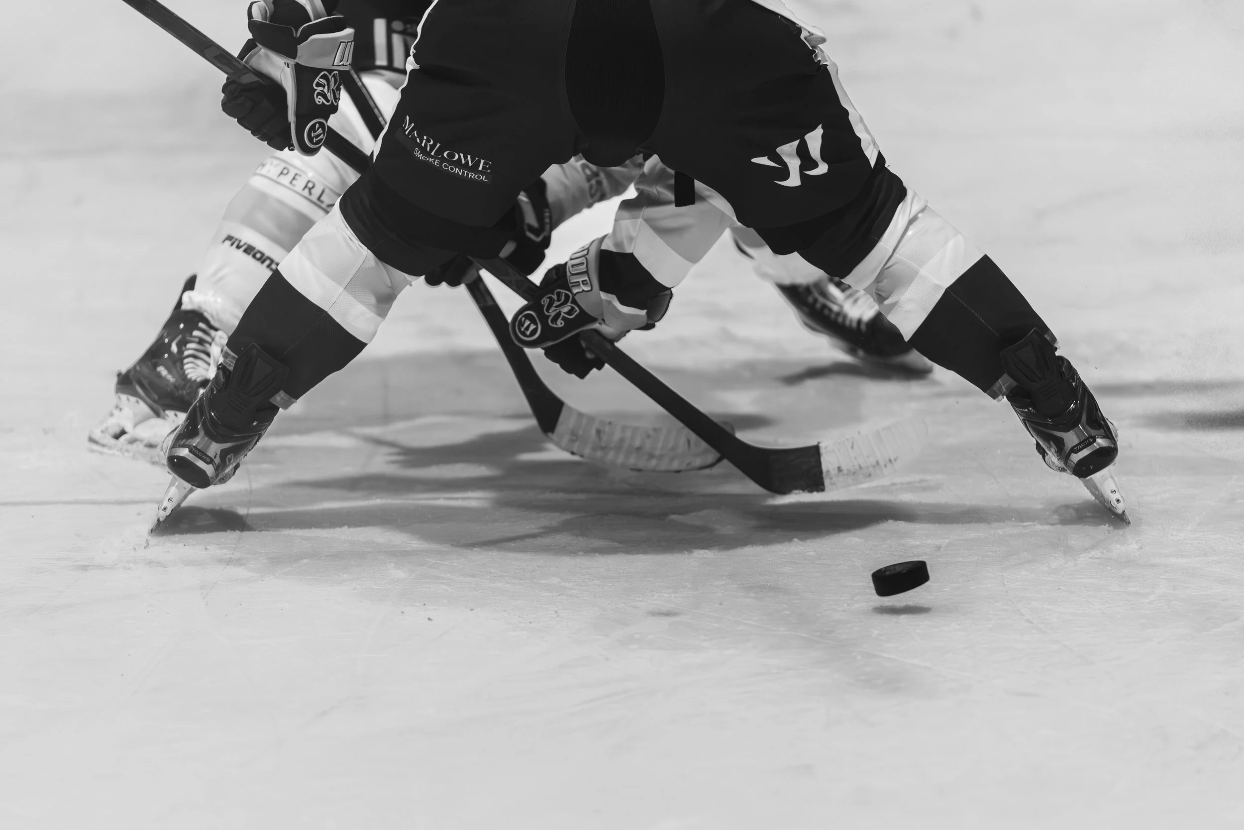 Two hockey players in a face-off, lower bodies bent, one with a stick on the ice, puck nearby, in an ice rink.