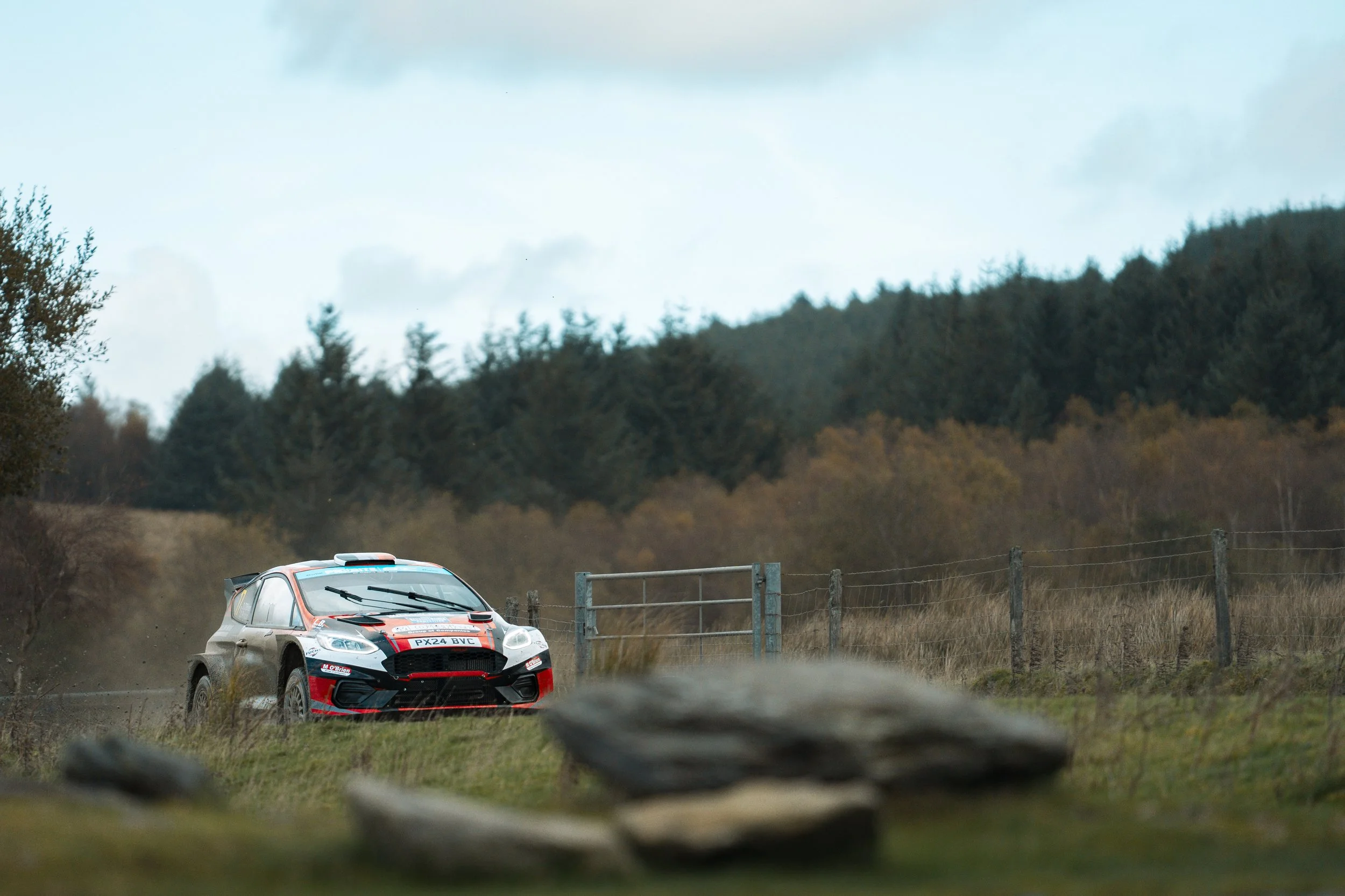 A rally car racing on a dirt road in a rural landscape with trees and hills in the background