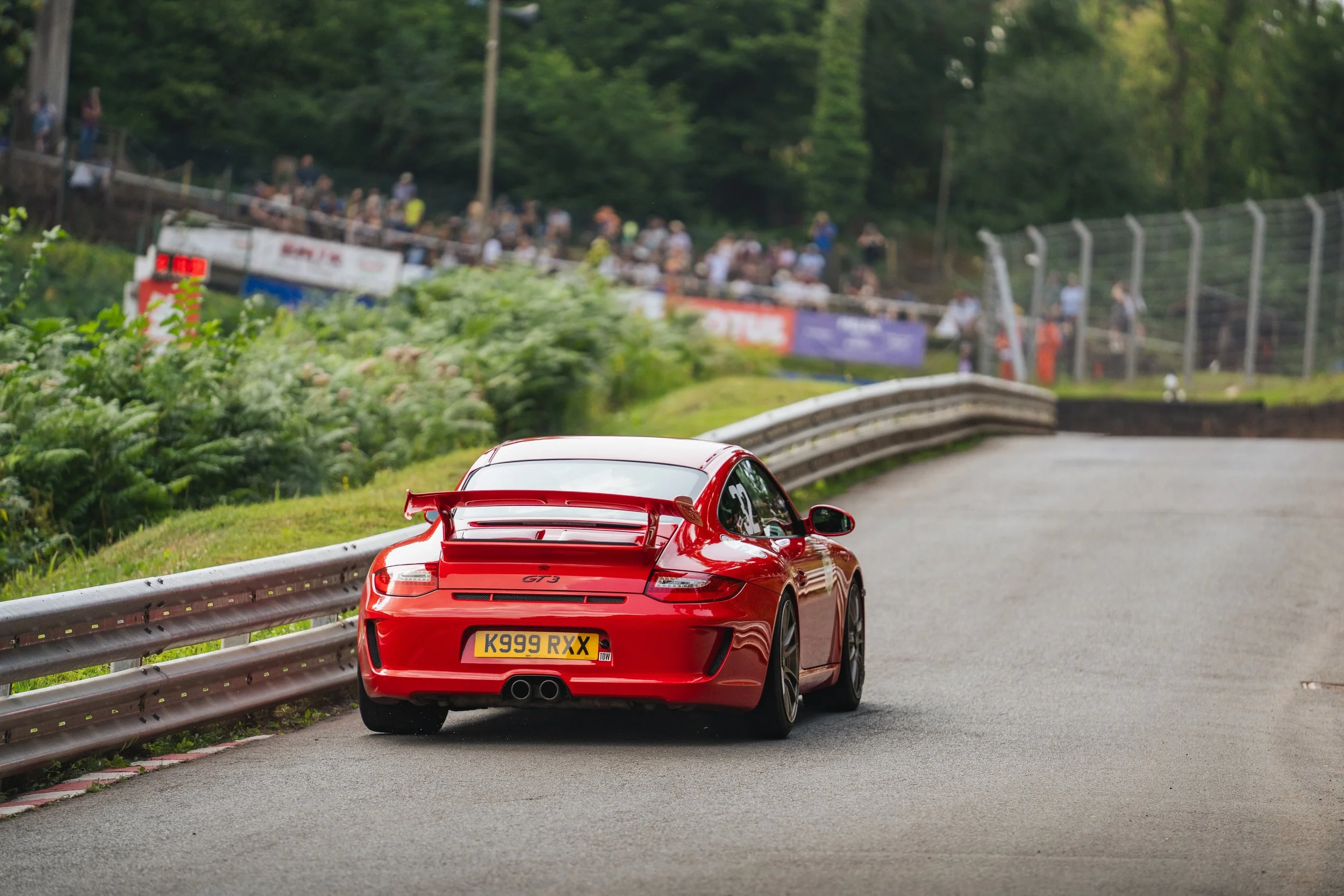 Red Porsche 911 GT3 racing car on a track, with spectators watching from the stands in the background.