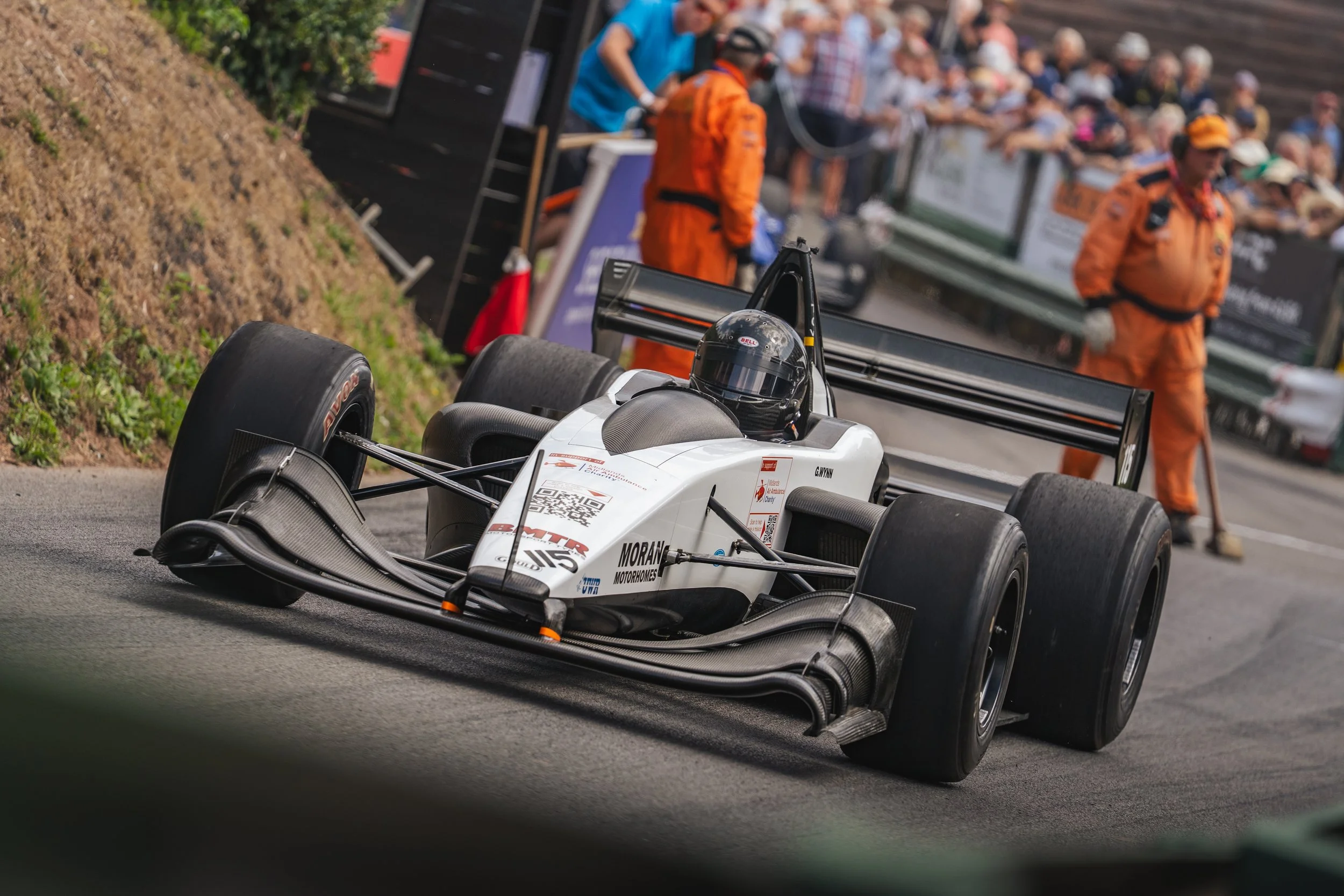 A white and black open-wheel race car on a track with a driver wearing a black helmet. In the background, people and officials in orange jumpsuits and casual clothing are observing, with some standing behind barriers.