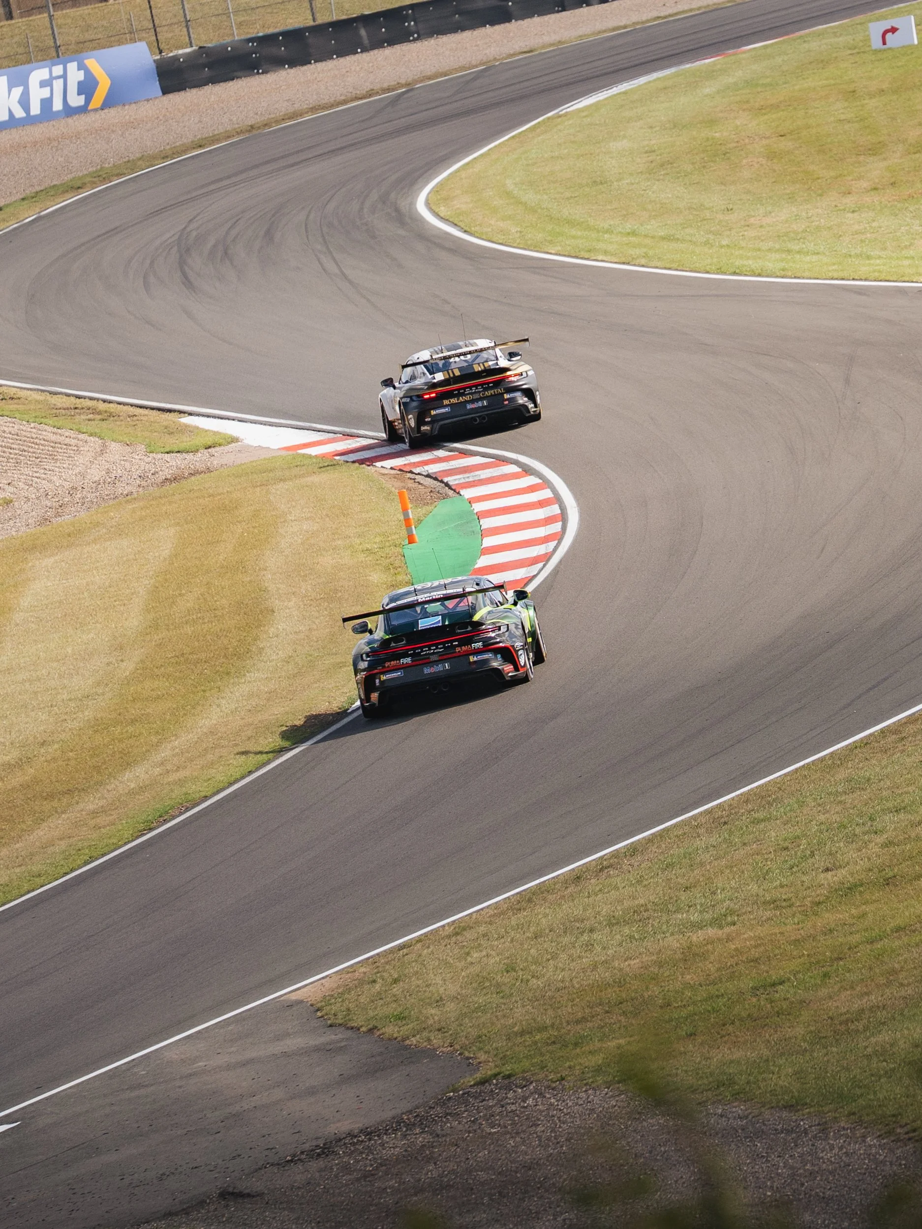 Two race cars navigating a turn on a racetrack, with one car slightly off the track onto the grass, during a motorsport event.