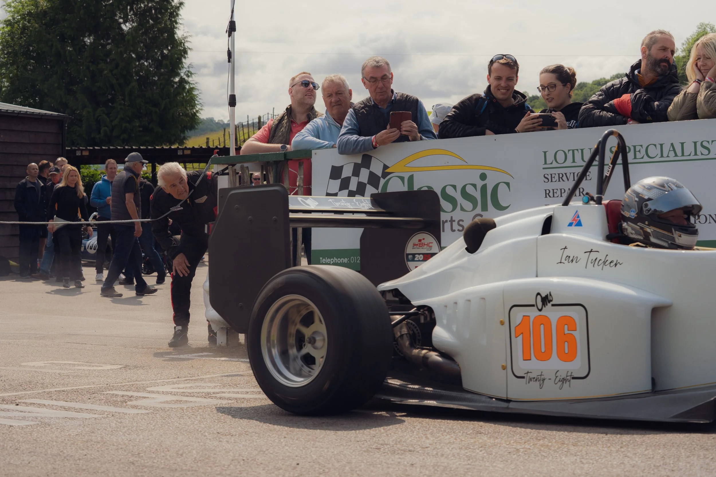 Race car with the number 106, called 'Twenty-Eight,' in a pit area with a driver helmet on the nose. A group of people are standing above on a platform, watching and taking pictures, while others stand in line behind the car.