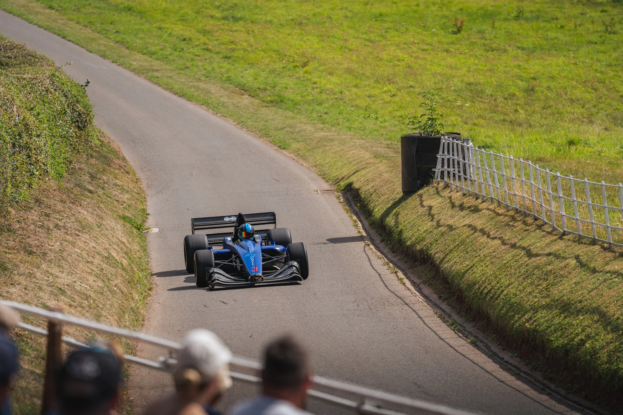 A blue race car with a driver wearing a helmet navigates a curved, grassy race track with spectators watching from the foreground.