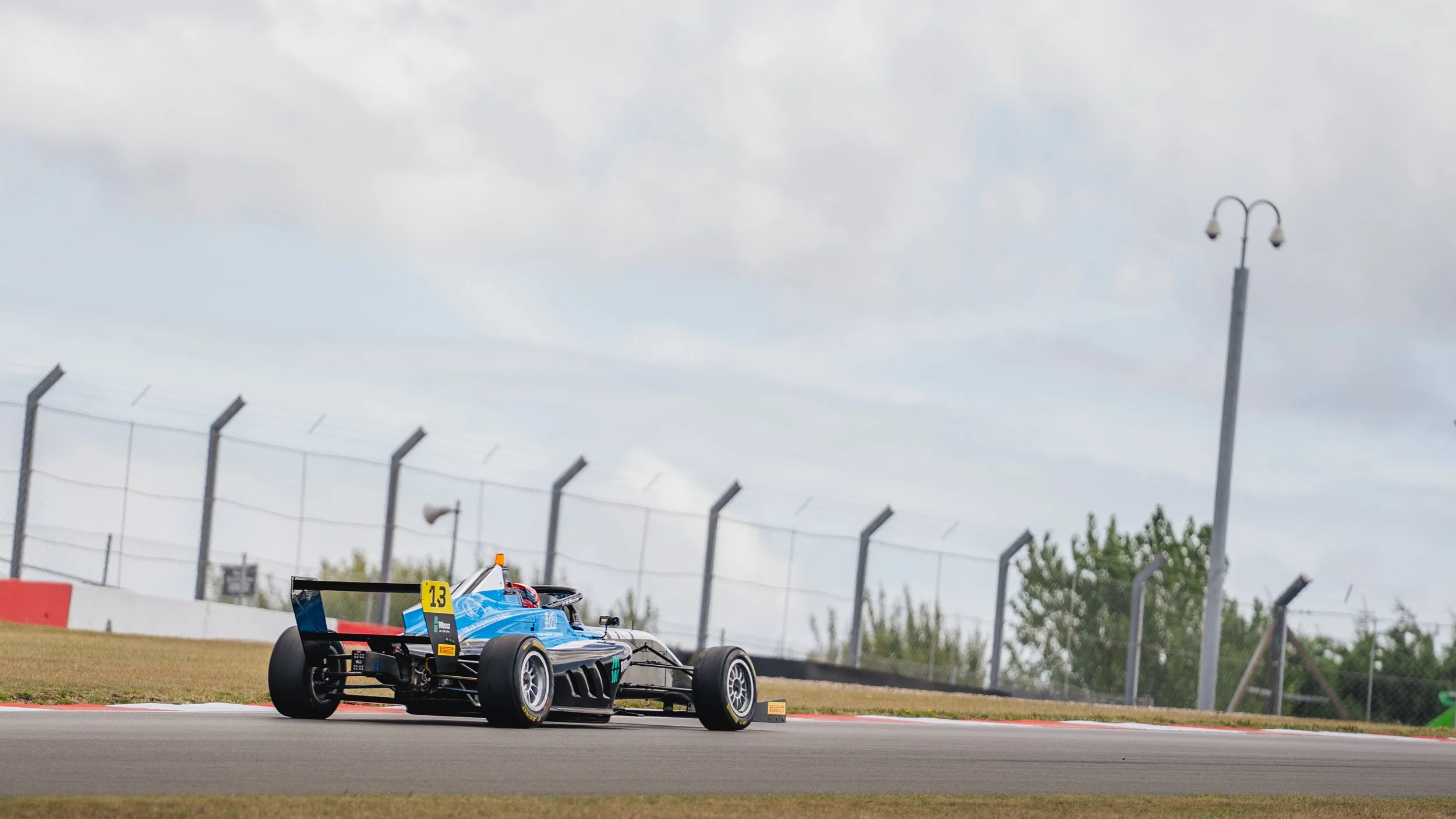 A blue race car on a race track with a safety barrier and a cloudy sky overhead.