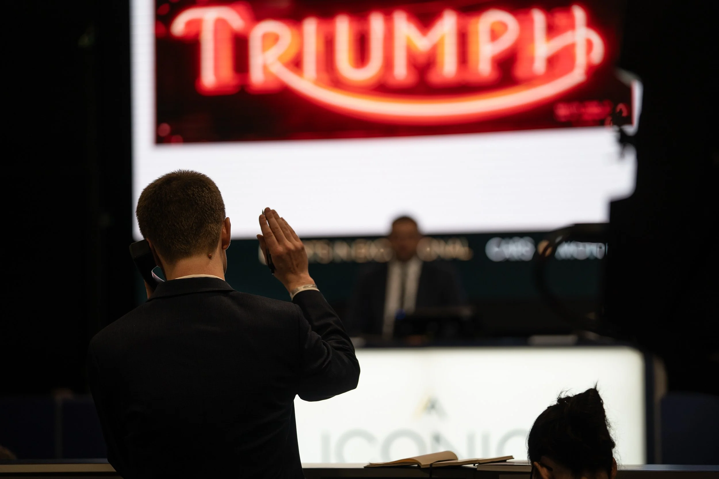 Auctioneer taking bids below a glowing Triumph neon sign at Race Retro