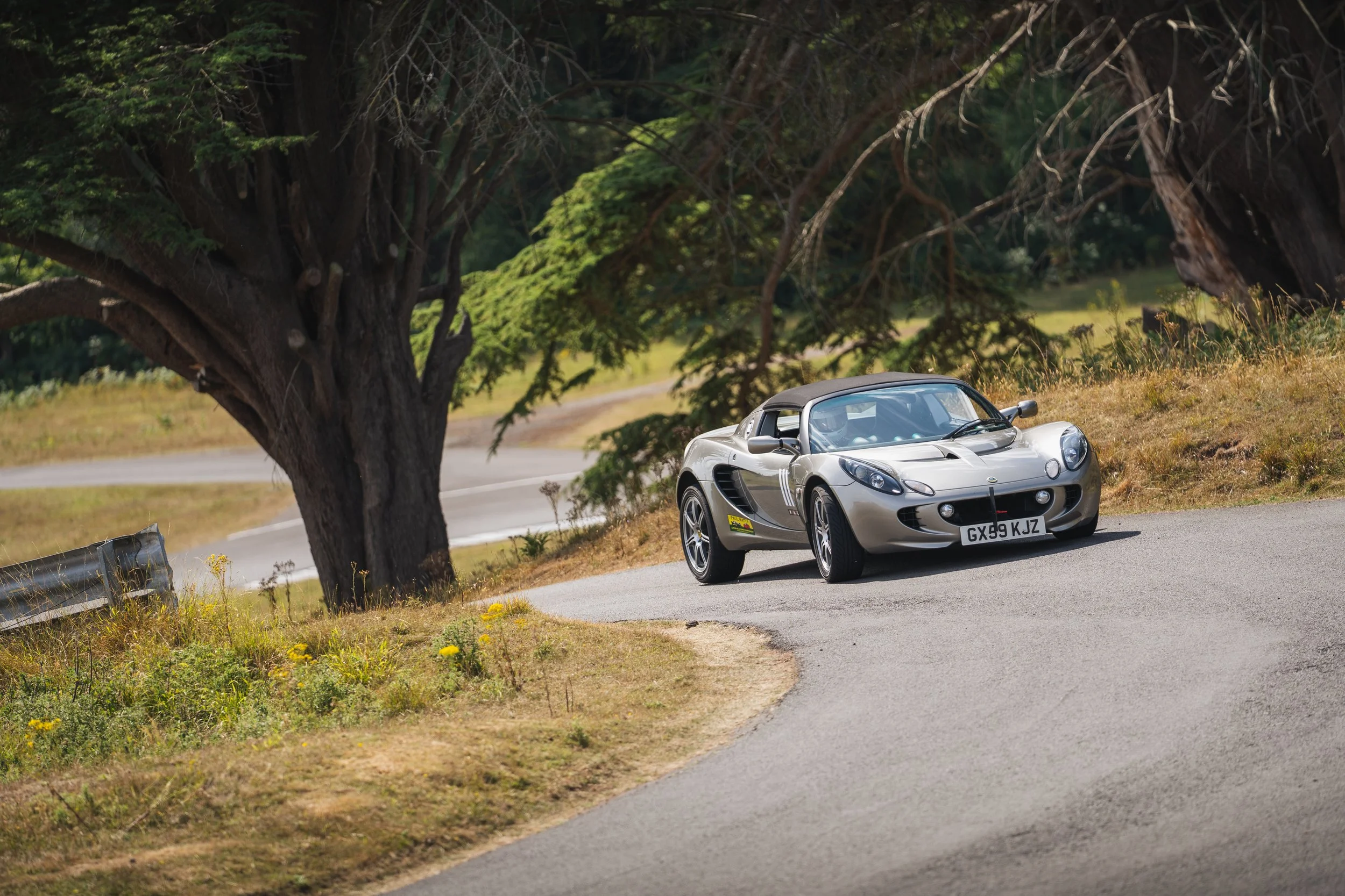 A silver Lotus Elise sports car with a black soft top driving on a winding countryside road, shaded by large trees.