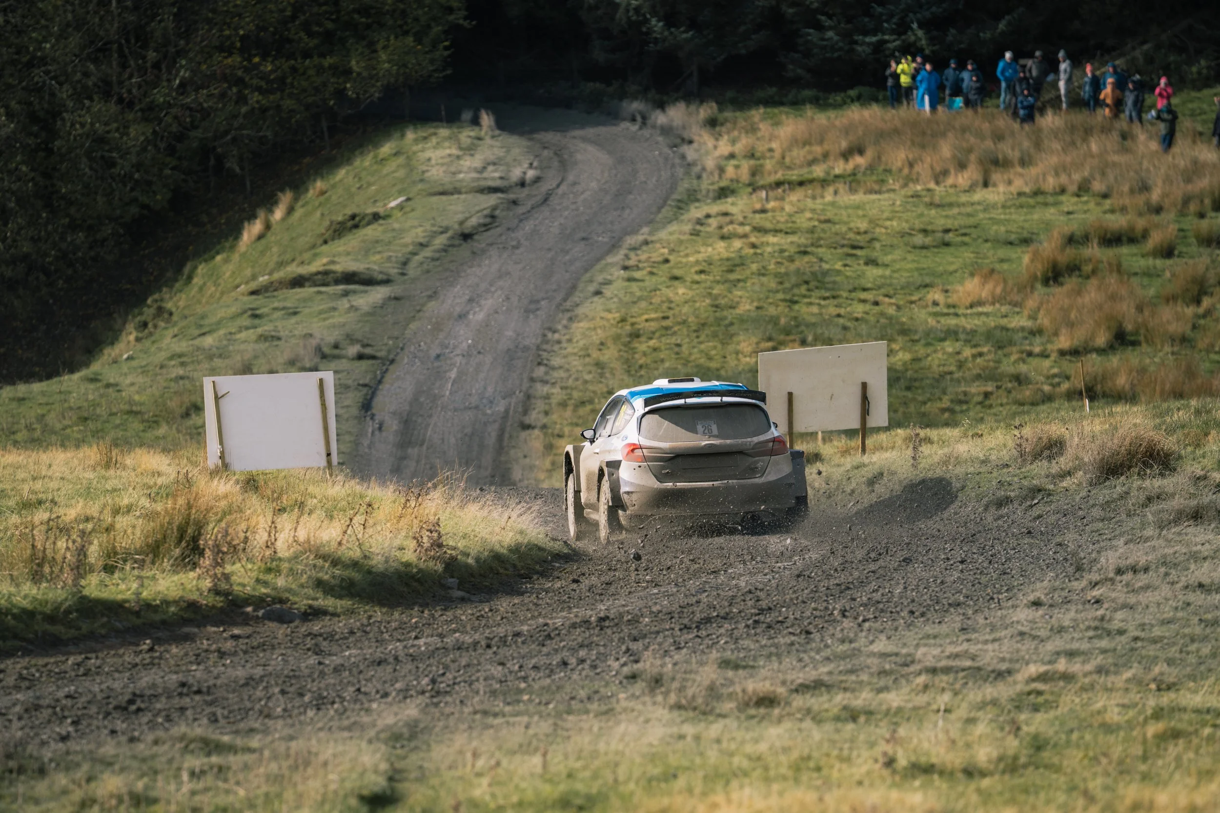 A rally car racing on a dirt road in a grassy, hilly landscape with spectators watching from the hillside.