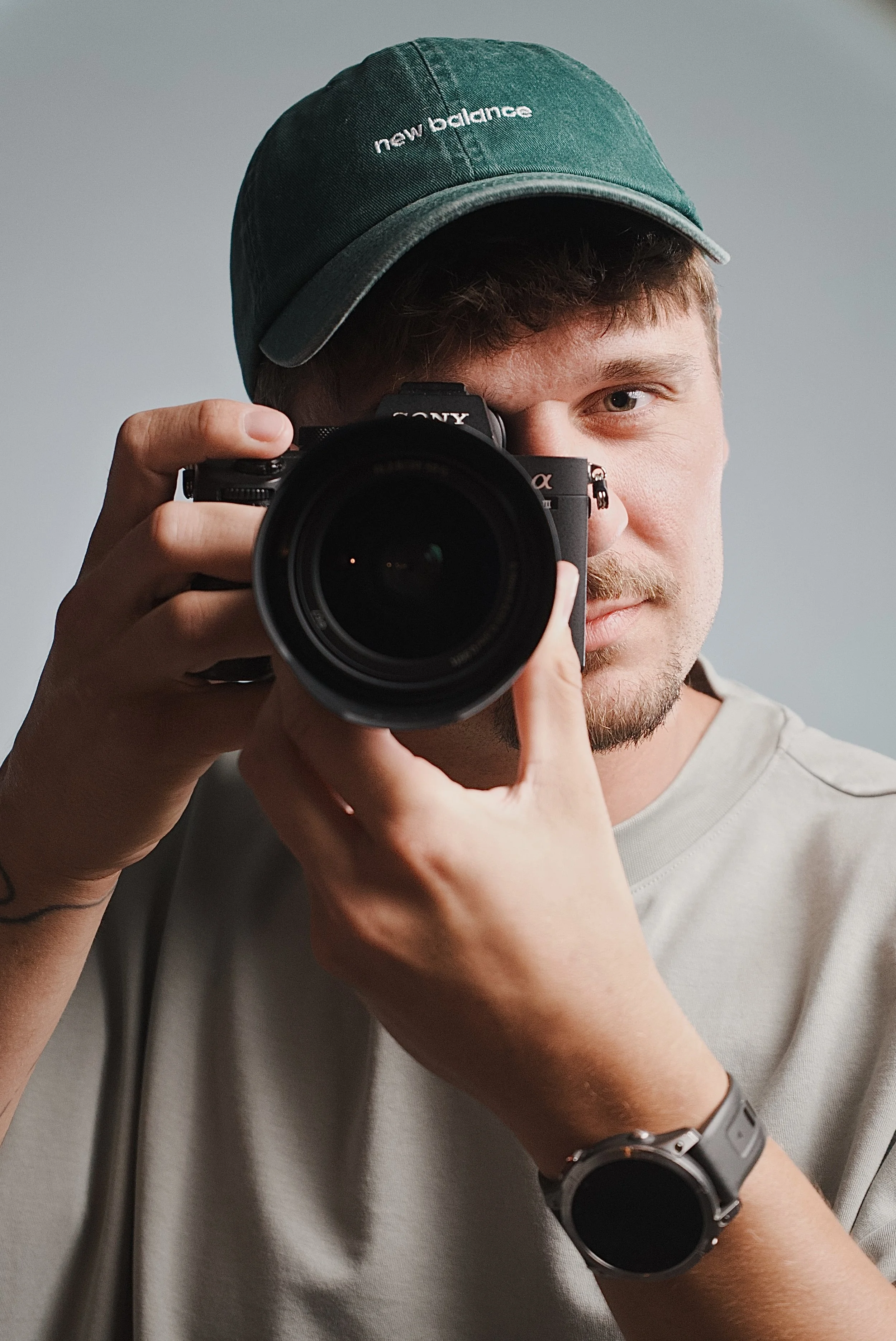 A young man with a beard and mustache, wearing a green New Balance baseball cap and a beige t-shirt, taking a selfie with a Sony mirrorless camera. He has a smartwatch on his left wrist and a bracelet on his right wrist, and is standing against a plain gray background.