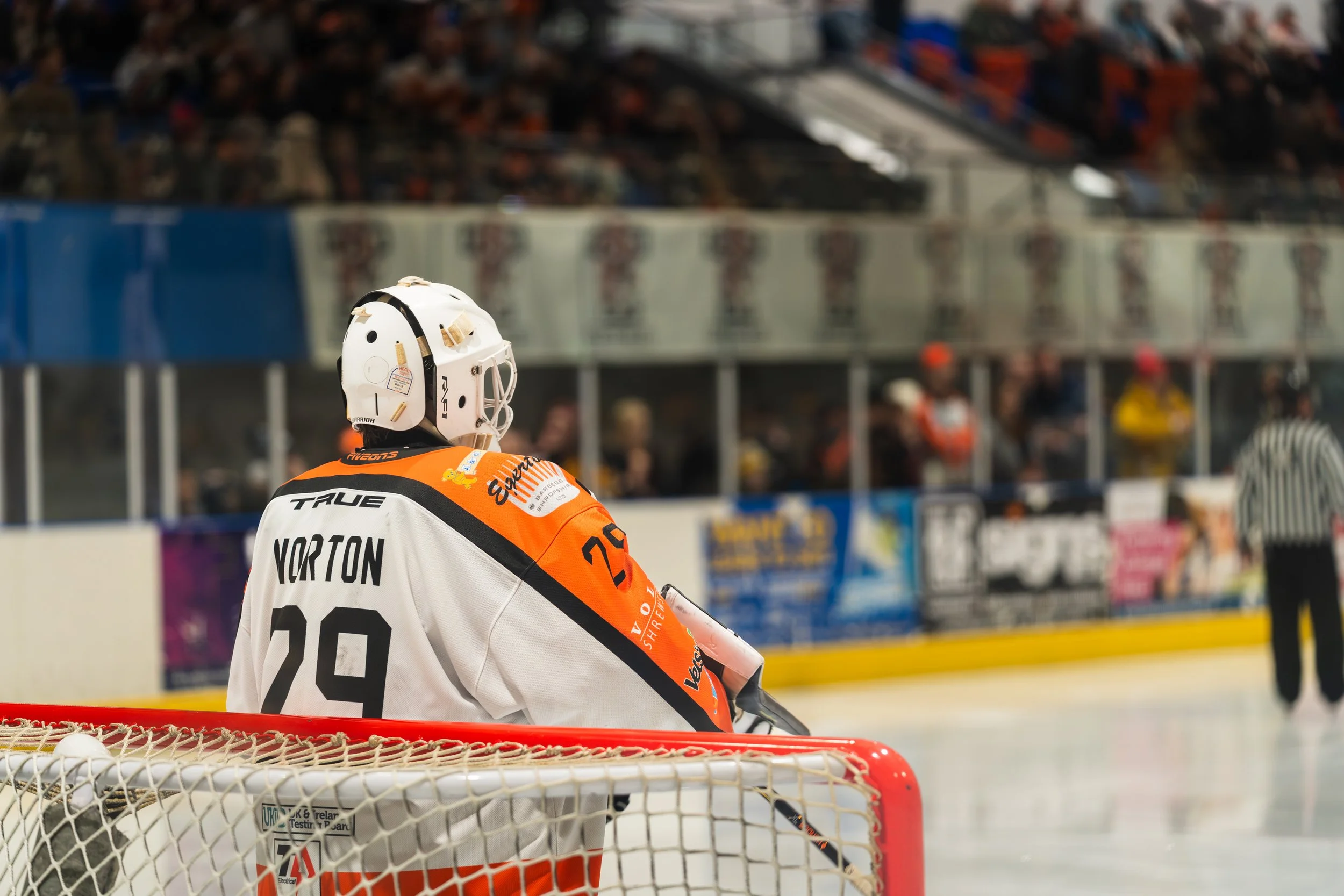 Hockey goalie in white, black, and orange uniform standing in front of the net during a game, with spectators in the background.