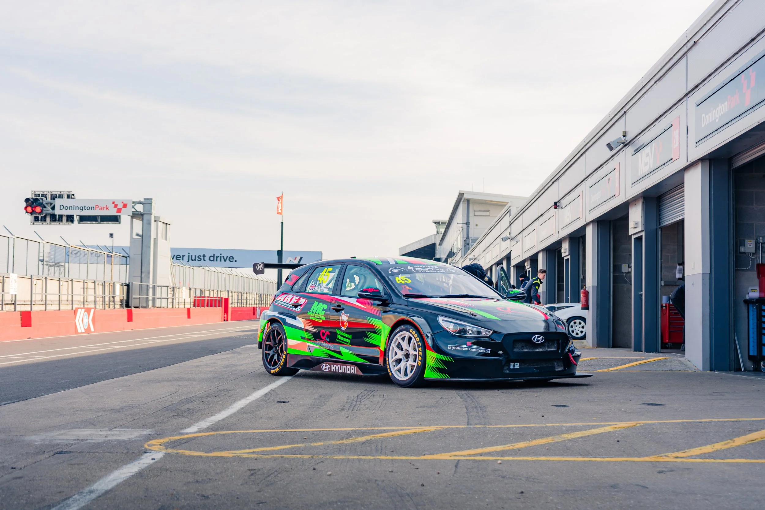 Race car in the pit lane at Donington Park race track with team members working on it and garages in the background.