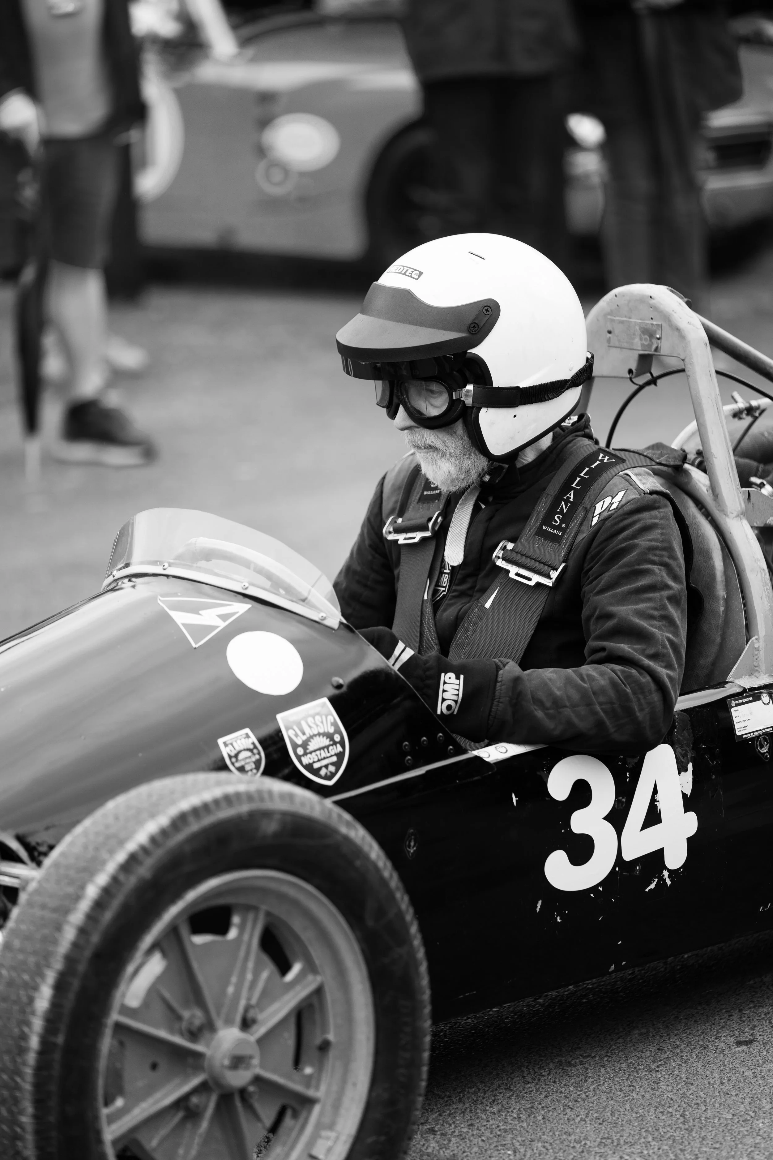 A man with a beard wearing a racing helmet, goggles, and racing gear sitting in a vintage race car with the number 34, preparing for a race.