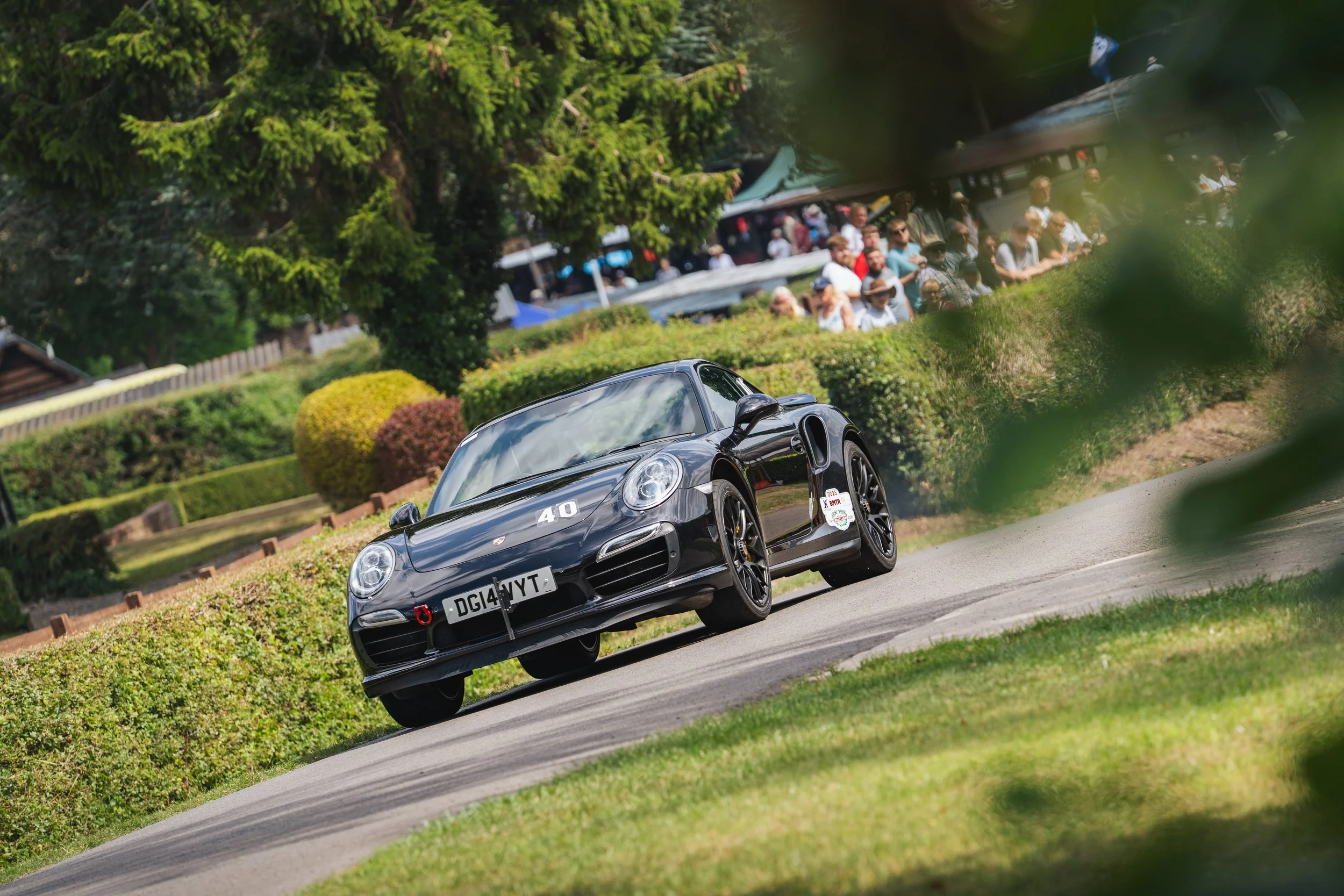 A black Porsche race car with the number 40 on the hood, driving on a curved road with a crowd of spectators in the background, surrounded by green trees and bushes.
