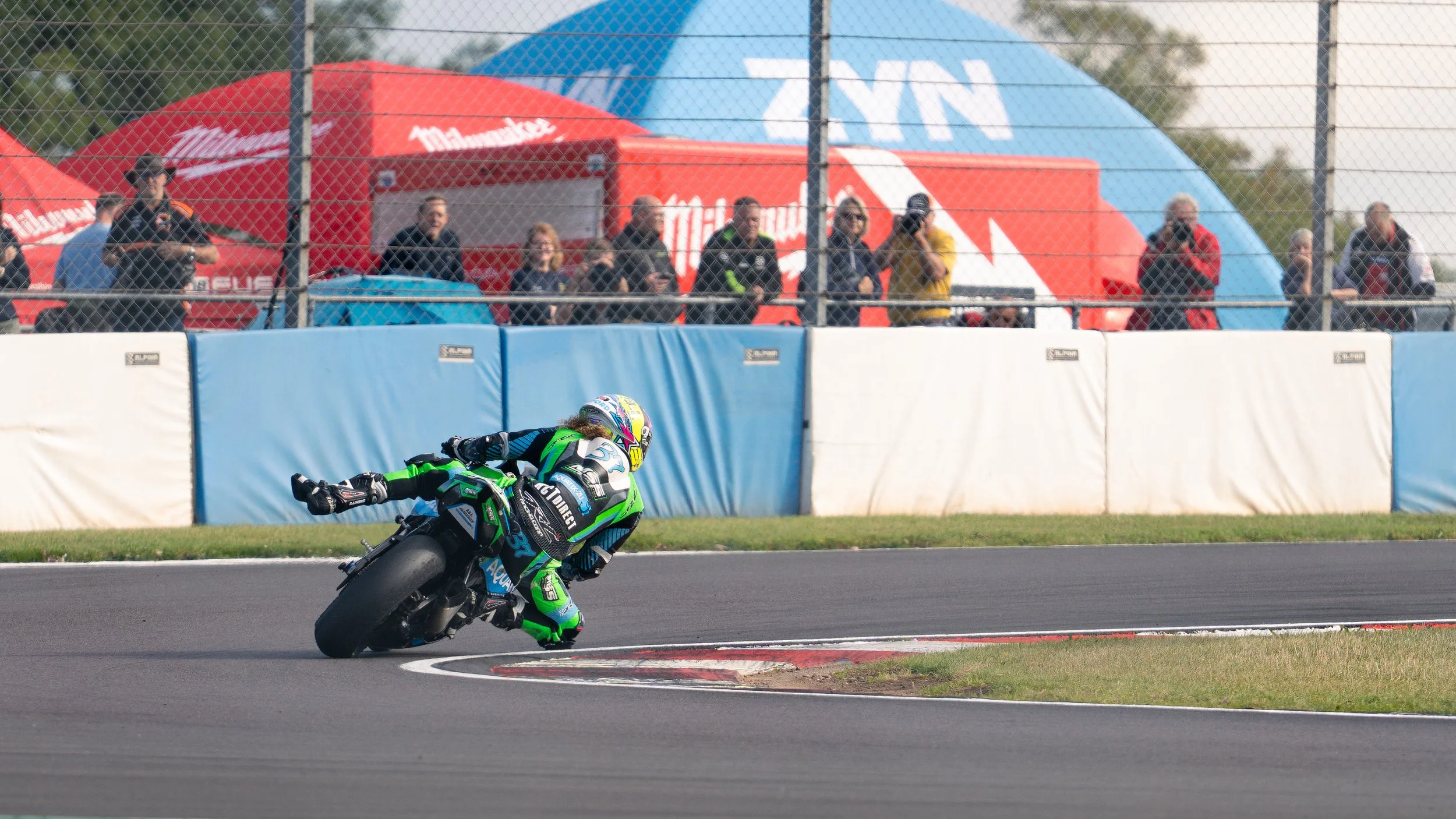 Motorcycle racing on a race track with spectators watching behind safety barriers and tents.
