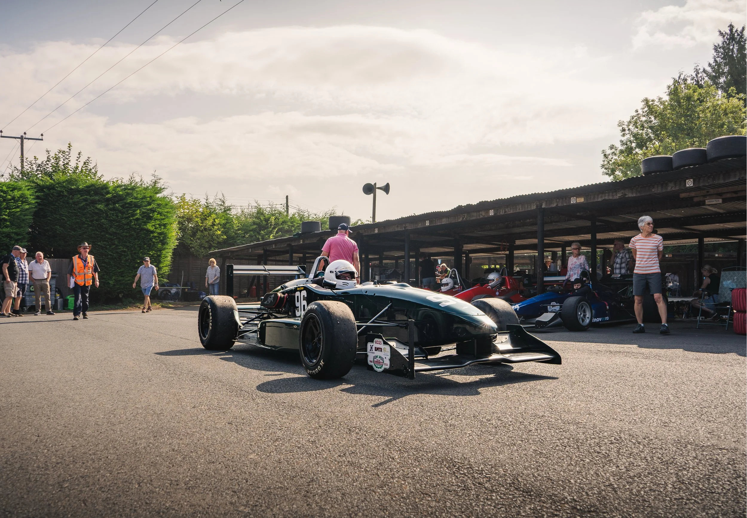 A vintage open-wheel race car, numbered 96, parked on a racetrack with a helmeted driver inside. Several people are standing and walking nearby, some under a covered area with more race cars. The scene is outdoors on a sunny day with trees and power 