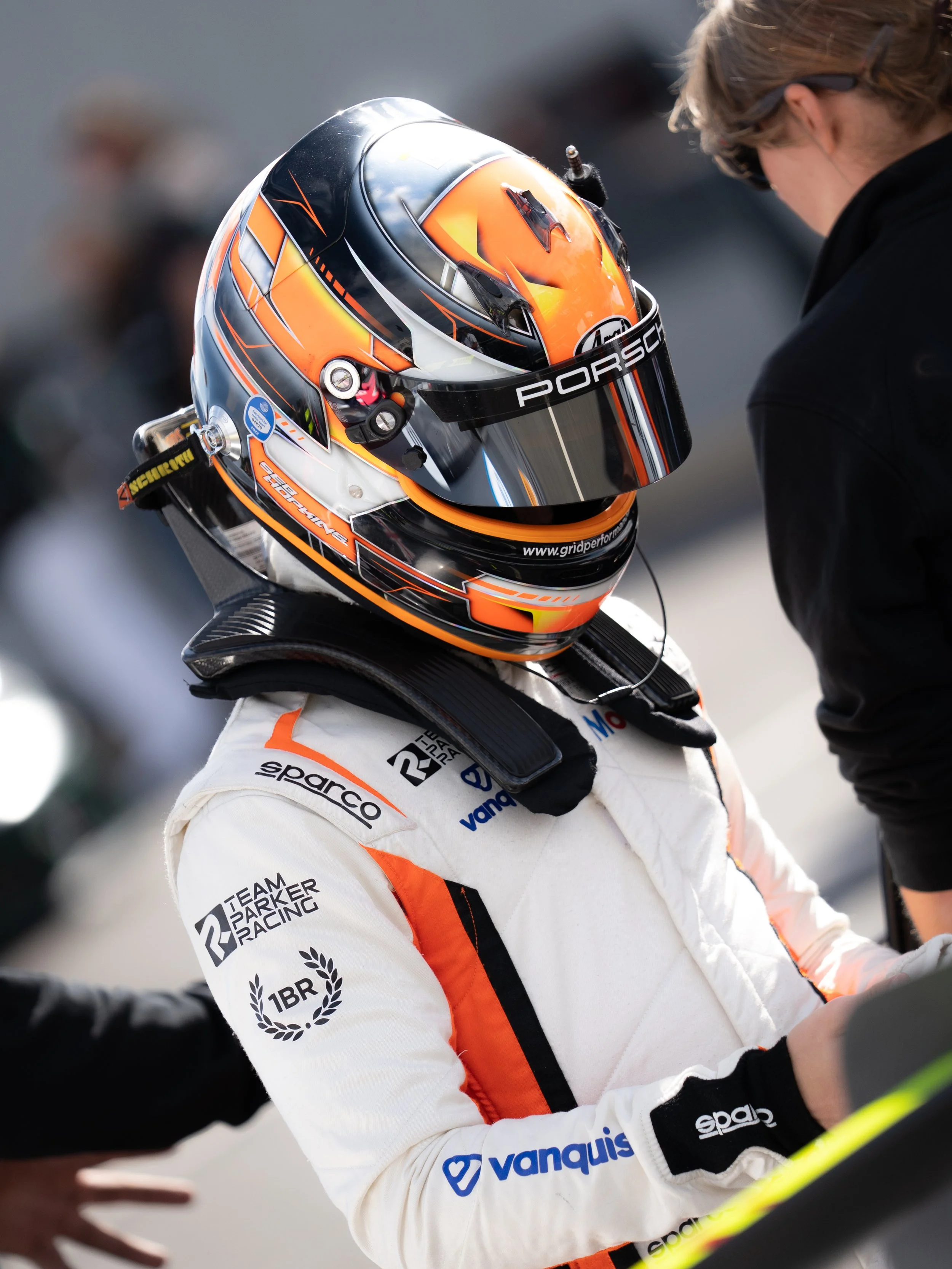 Race car driver in a racing suit and helmet in discussion with another person.