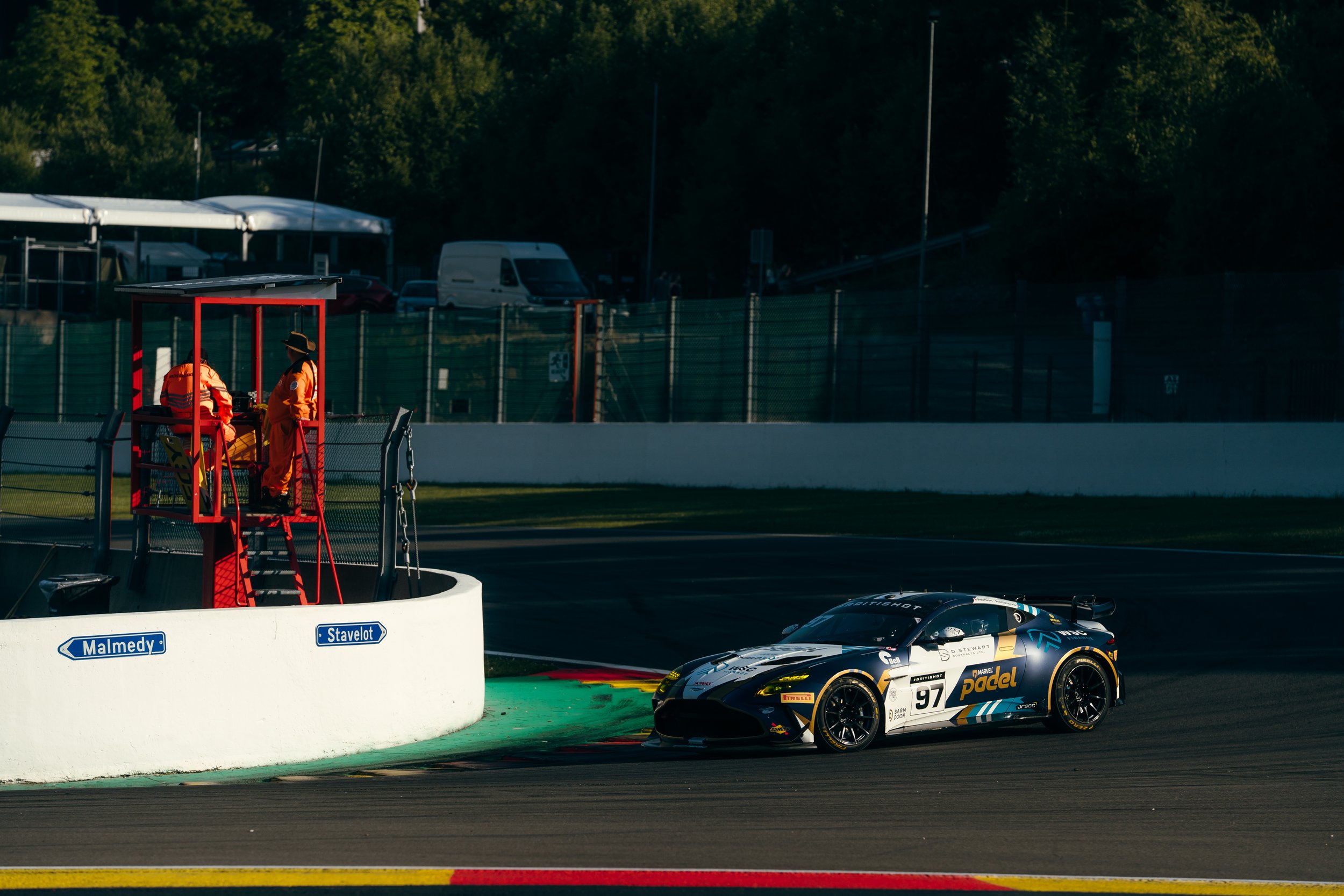 A race car with the number 97 on it is on a racetrack near the pit lane. Two officials in orange uniforms are in a raised observation booth above the track. There is a green and black painted curb at the edge of the track, and a white wall barrier se