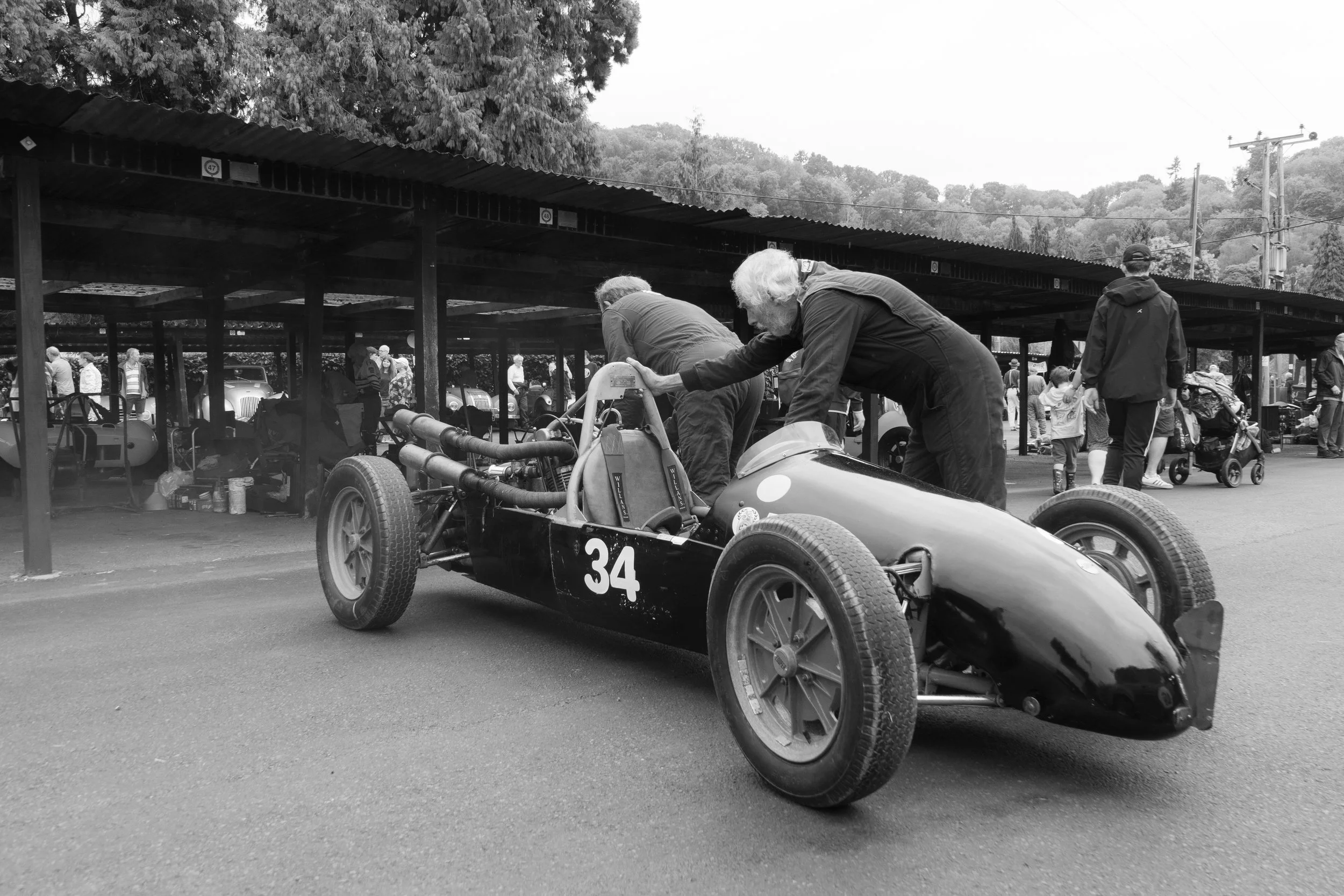 Black and white photo of vintage race car with the number 34 on its side, being prepared by two older men at a car show or race event, with people and classic cars in the background.