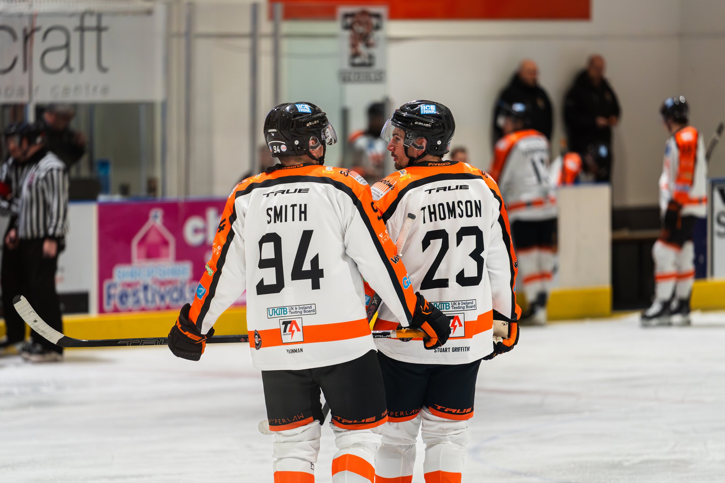 Two ice hockey players in white and orange jerseys, number 94 and 23, standing on the ice, with others in the background near the rink's boards.