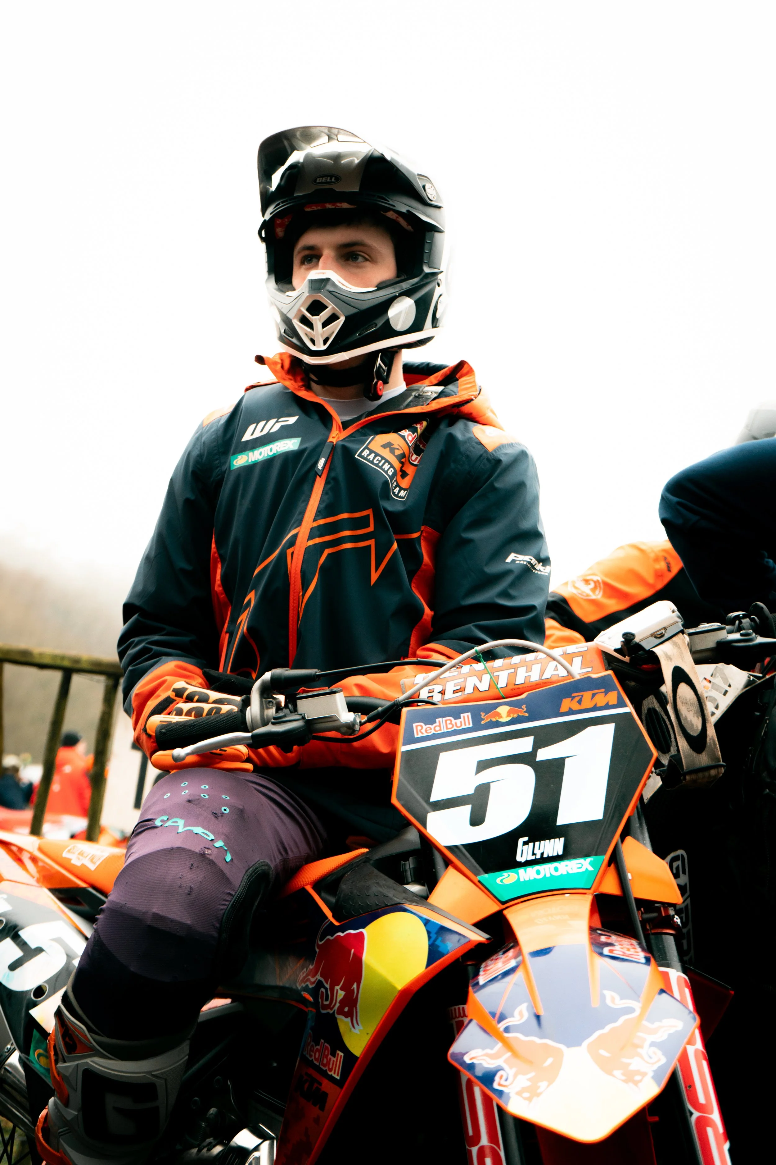 A motorcyclist in racing gear and helmet sitting on an orange KTM motorcycle with racing decals and the number 51. The background is cloudy and outdoors.
