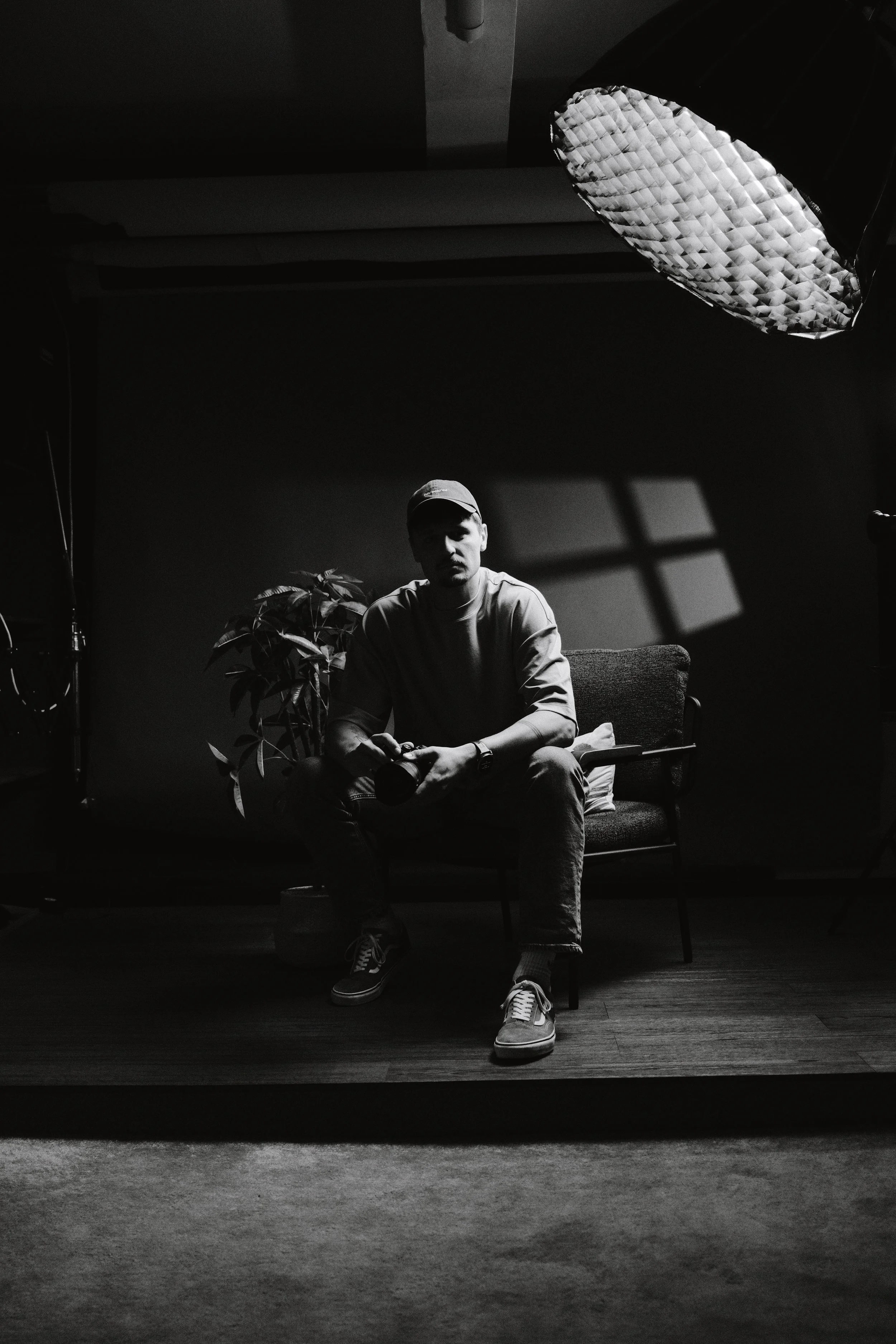 A man sitting on a chair in a dark room with a large umbrella reflector overhead. He is wearing a cap, t-shirt, jeans, and sneakers, and appears to be holding a camera. There is a plant behind him and a window or light source casting shadows on the wall.