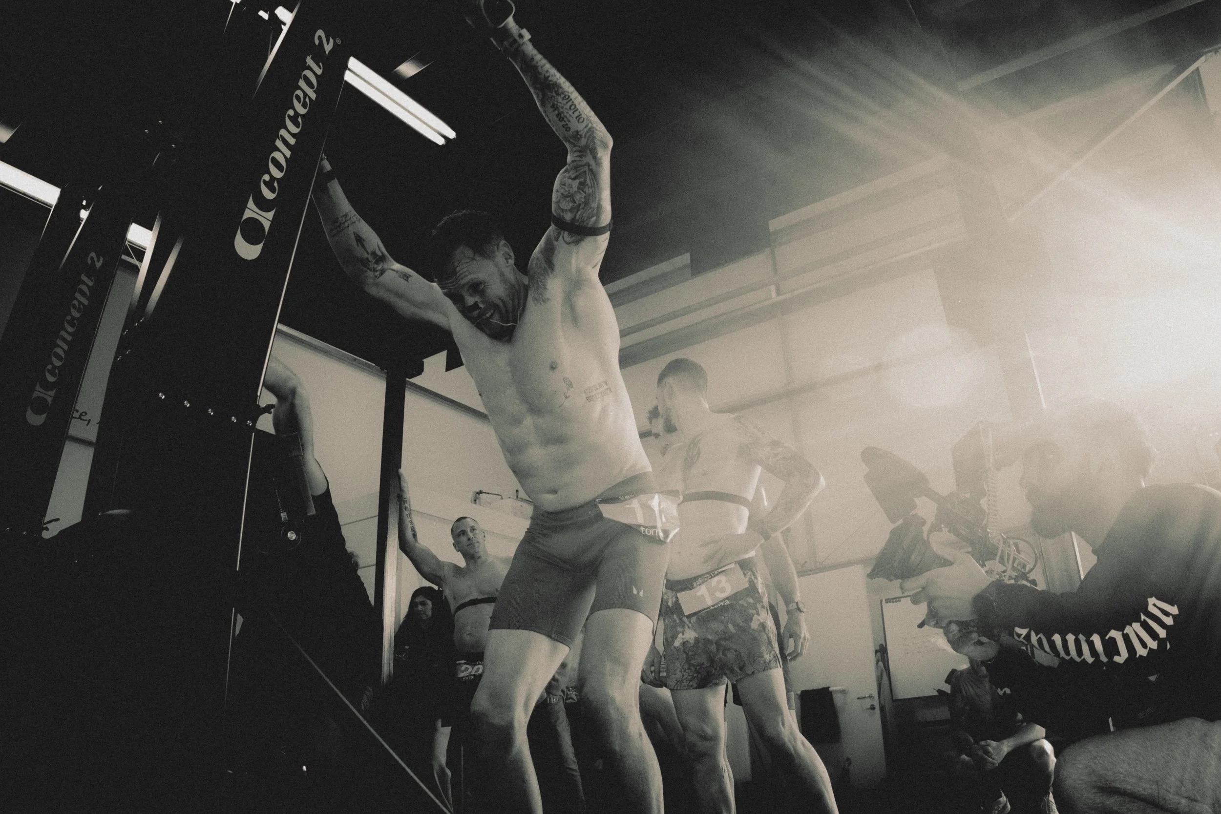 A shirtless man with tattoos and a black arm band is holding onto gym equipment with both hands, appearing to be in a gym or fitness environment. Other people are visible in the background, some standing and some engaging in fitness activities.