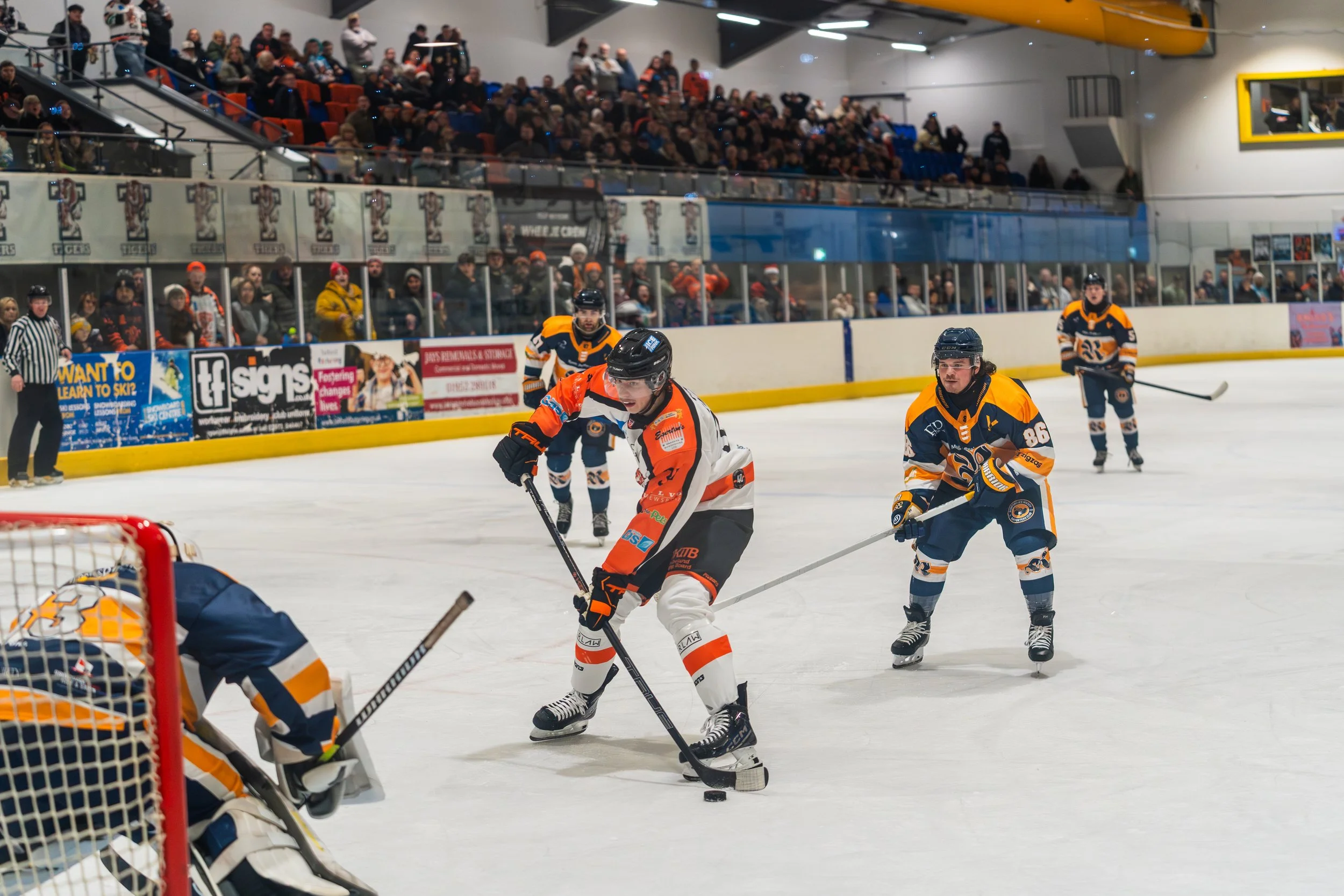 Hockey players on the ice rink during a game, with a crowd of spectators watching from the stands.