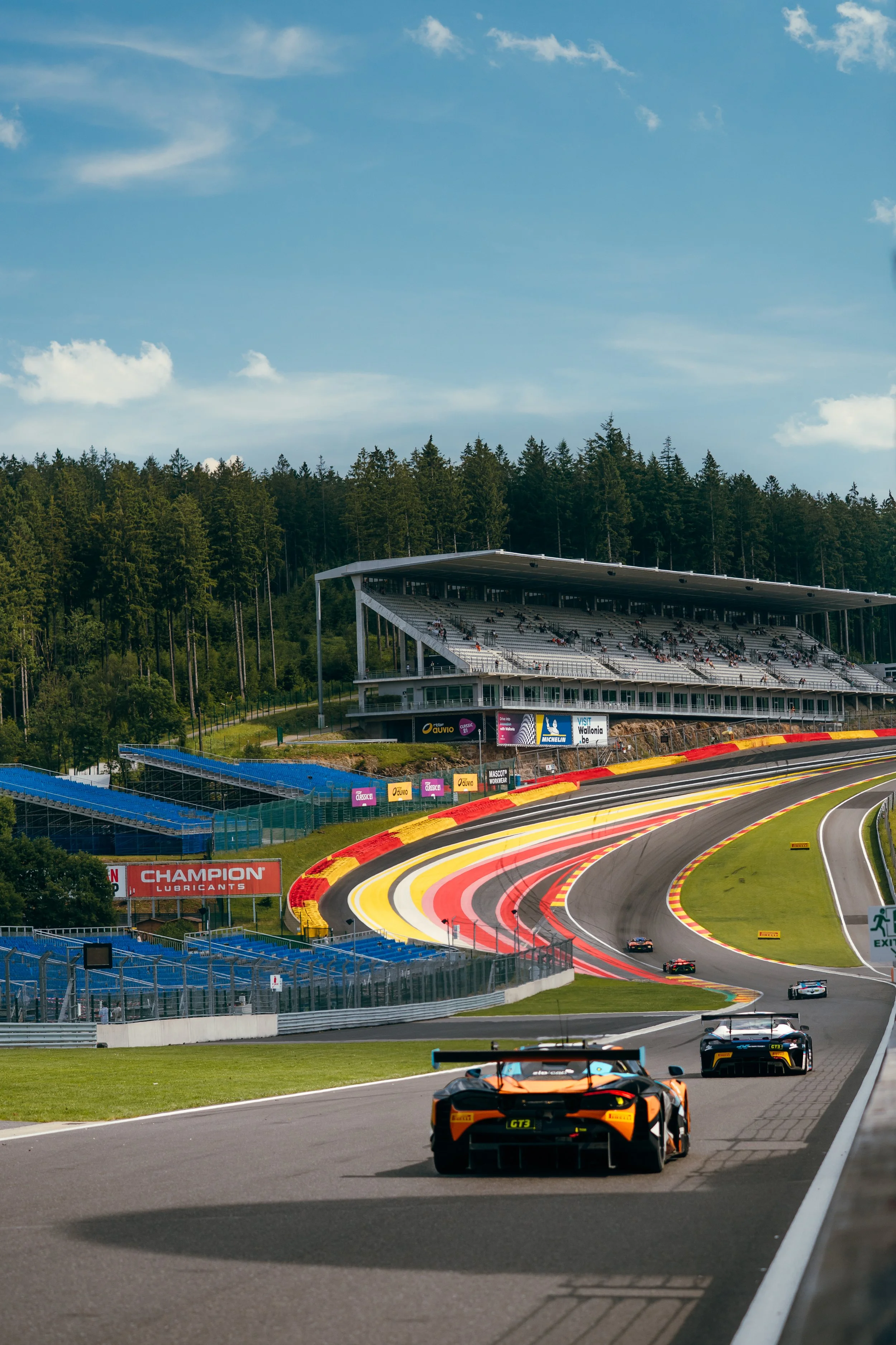 Race cars on a track with colorful curbing, a grandstand in the background, and a hillside covered with trees.